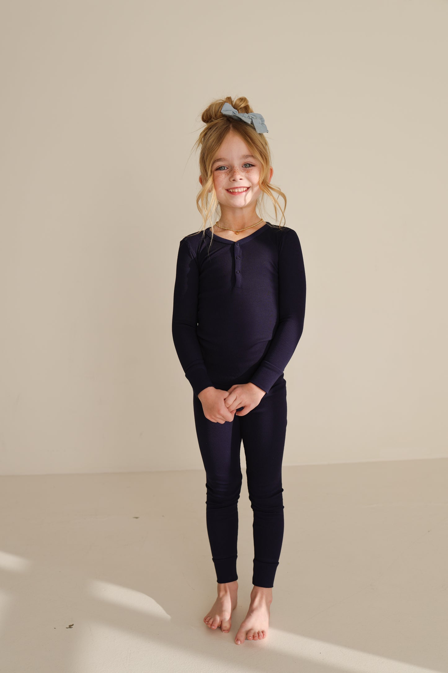 A young girl with long blonde hair stands barefoot on a light floor against a beige background, smiling in forever french baby’s Bamboo Two Piece Pajamas | Blueberry Ribbed.