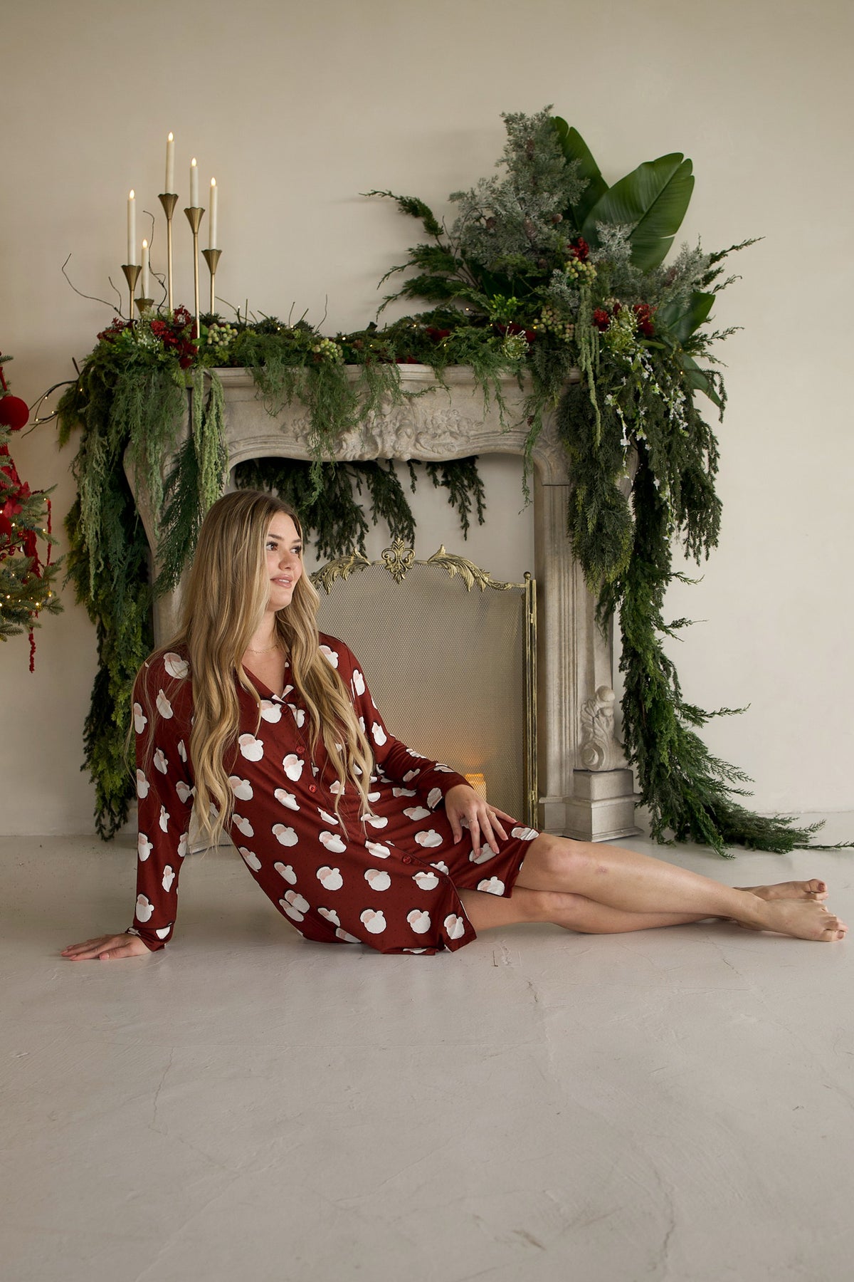 A woman wearing a forever french baby Women’s Bamboo Sleeping Dress | Vintage Santa sits on the floor in front of a fireplace decorated with greenery.