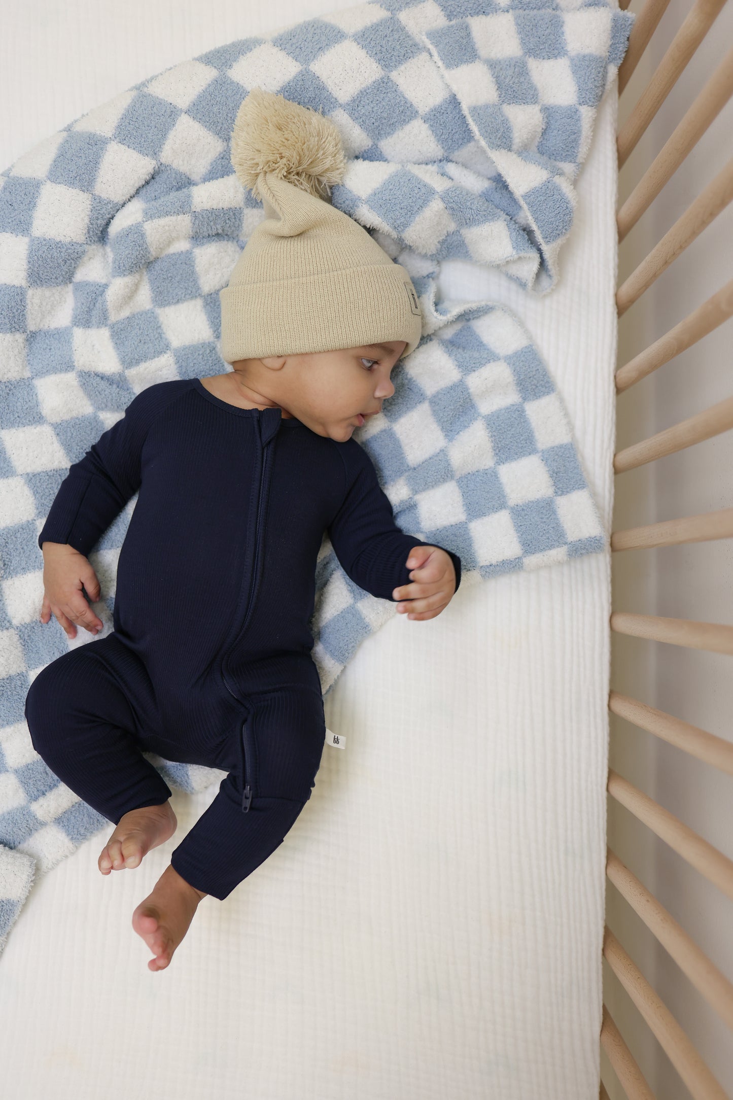 A baby in a beige knit hat with pom-poms and a navy blue onesie lies on the forever french baby Plush Blanket | Morning Check, atop a blue and white checkered crib mattress, surrounded by light wooden bars.