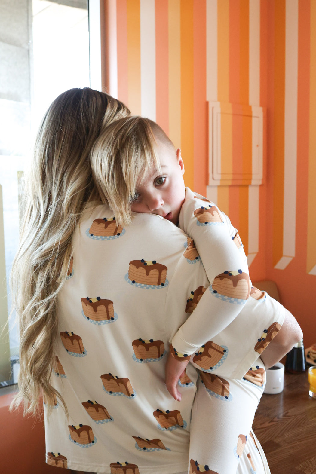 A woman with long blonde hair holds a young child in matching forever french baby Bamboo Two Piece Pajamas | Sunday Stack, featuring a pancake print. The child looks at the camera while resting on her shoulder against orange and yellow striped walls.