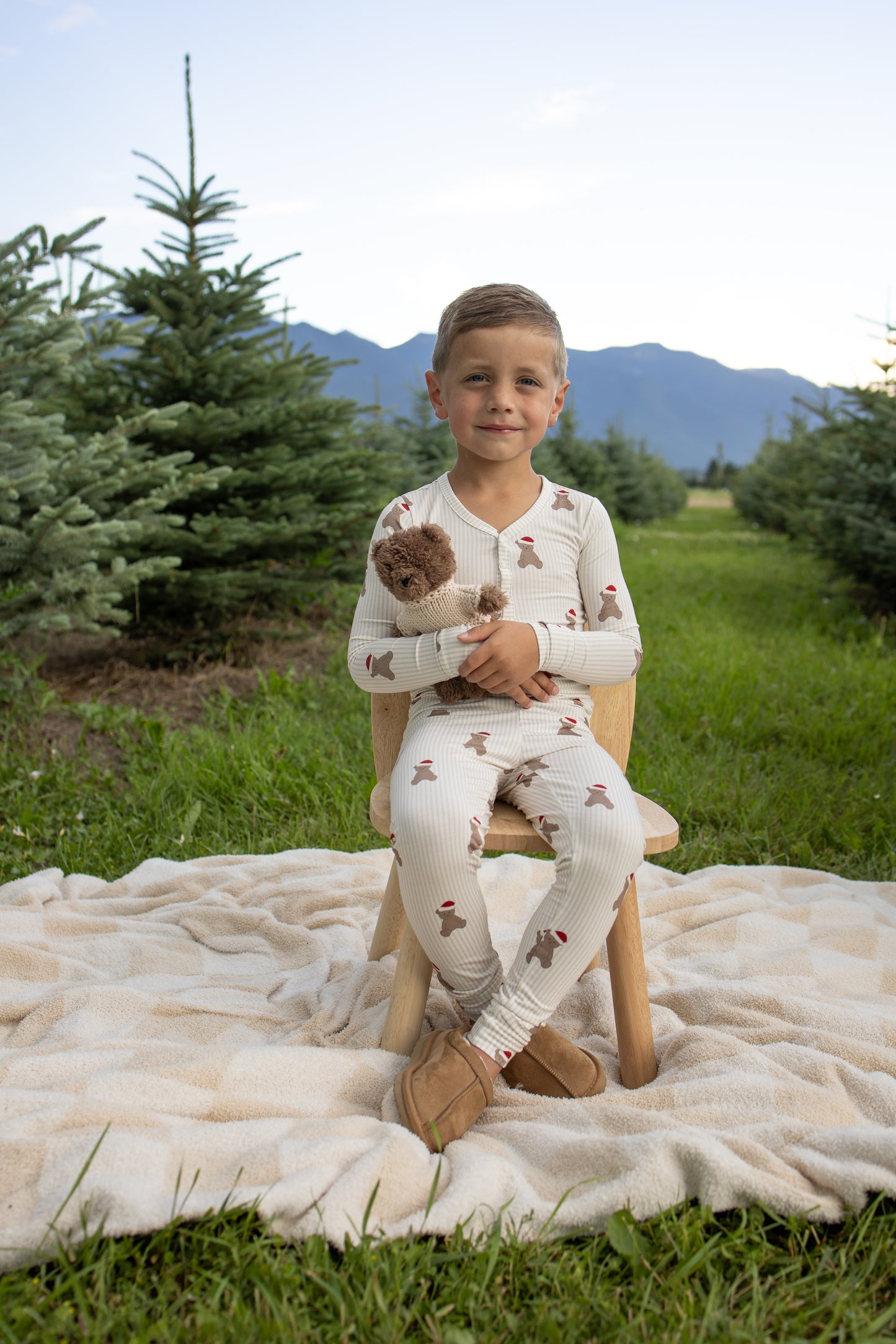 A young child wearing forever french baby's Bamboo Two Piece Pajamas in the Santa Teddy print sits on a wooden chair outdoors, cuddling a matching Santa Teddy. They are on a blanket between rows of evergreen trees with mountains in the background.