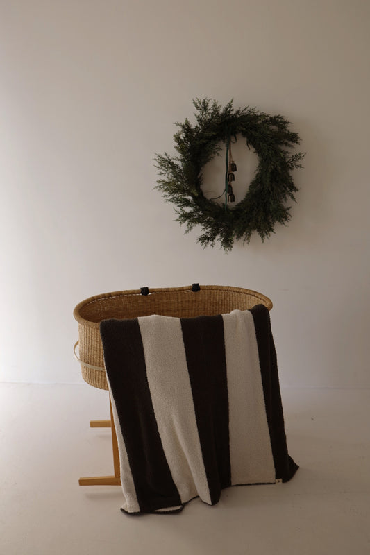 A woven bassinet with a forever french baby Plush Blanket | Fireside Stripe draped over the side sits on a wooden stand. A green wreath and black bells hang on the plain white wall above.