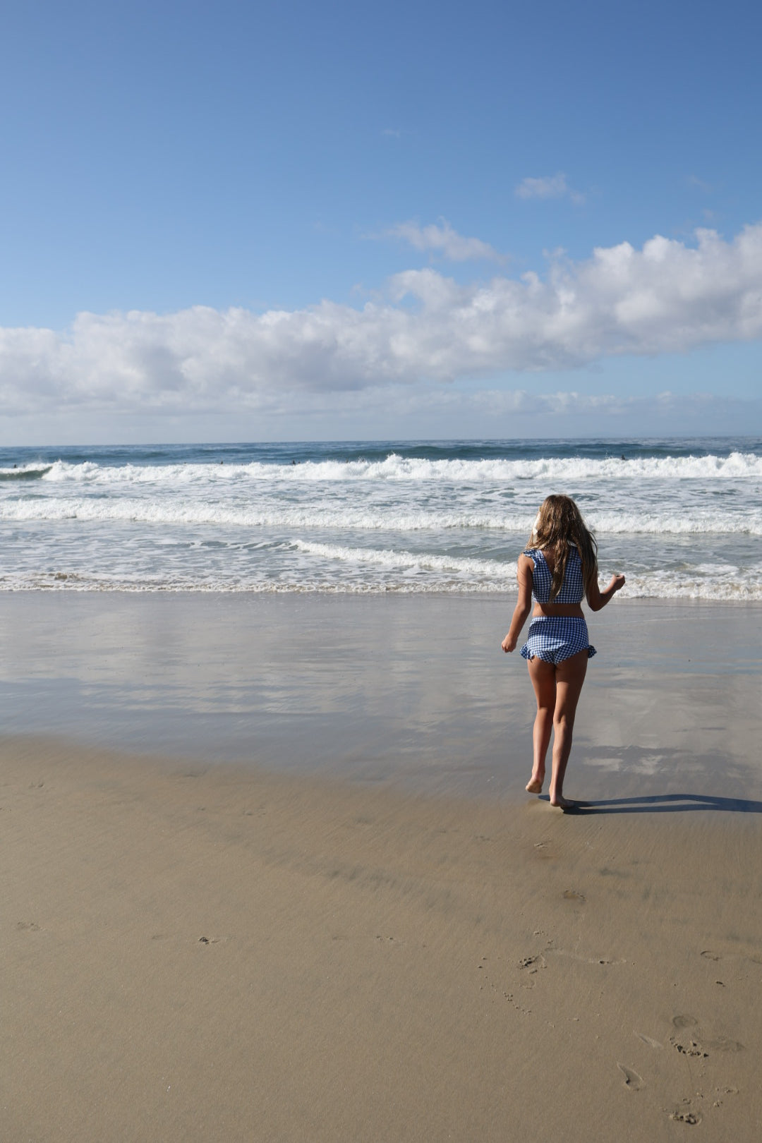 A young girl wears the forever french baby Girls Two Piece Swimsuit in Blue Gingham as she stands on a sandy beach, gazing at the ocean waves beneath a blue sky with scattered clouds.