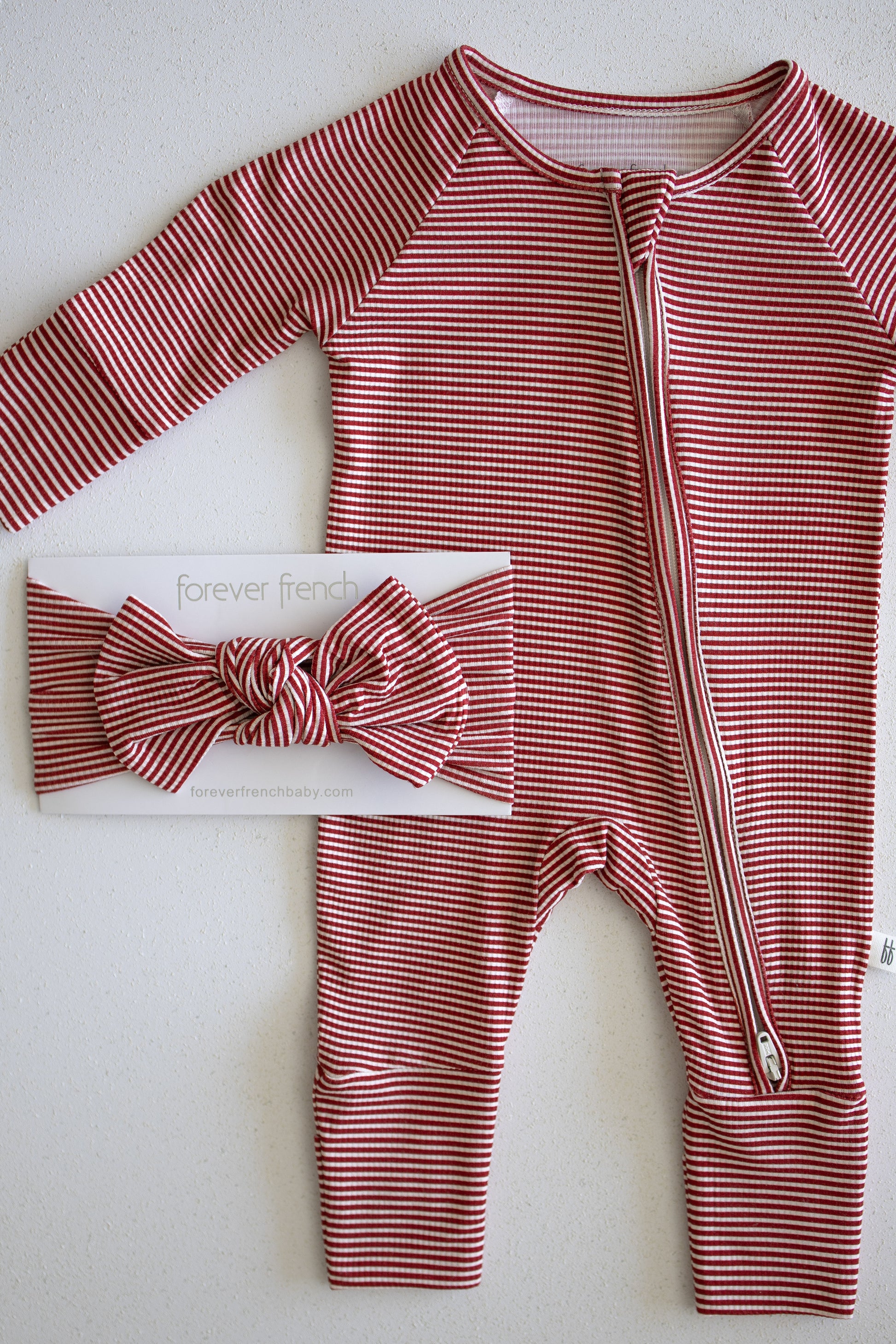 A red and white peppermint stripe bamboo head wrap from forever french baby is displayed alongside a matching zippered baby onesie, both laid out on a light surface.