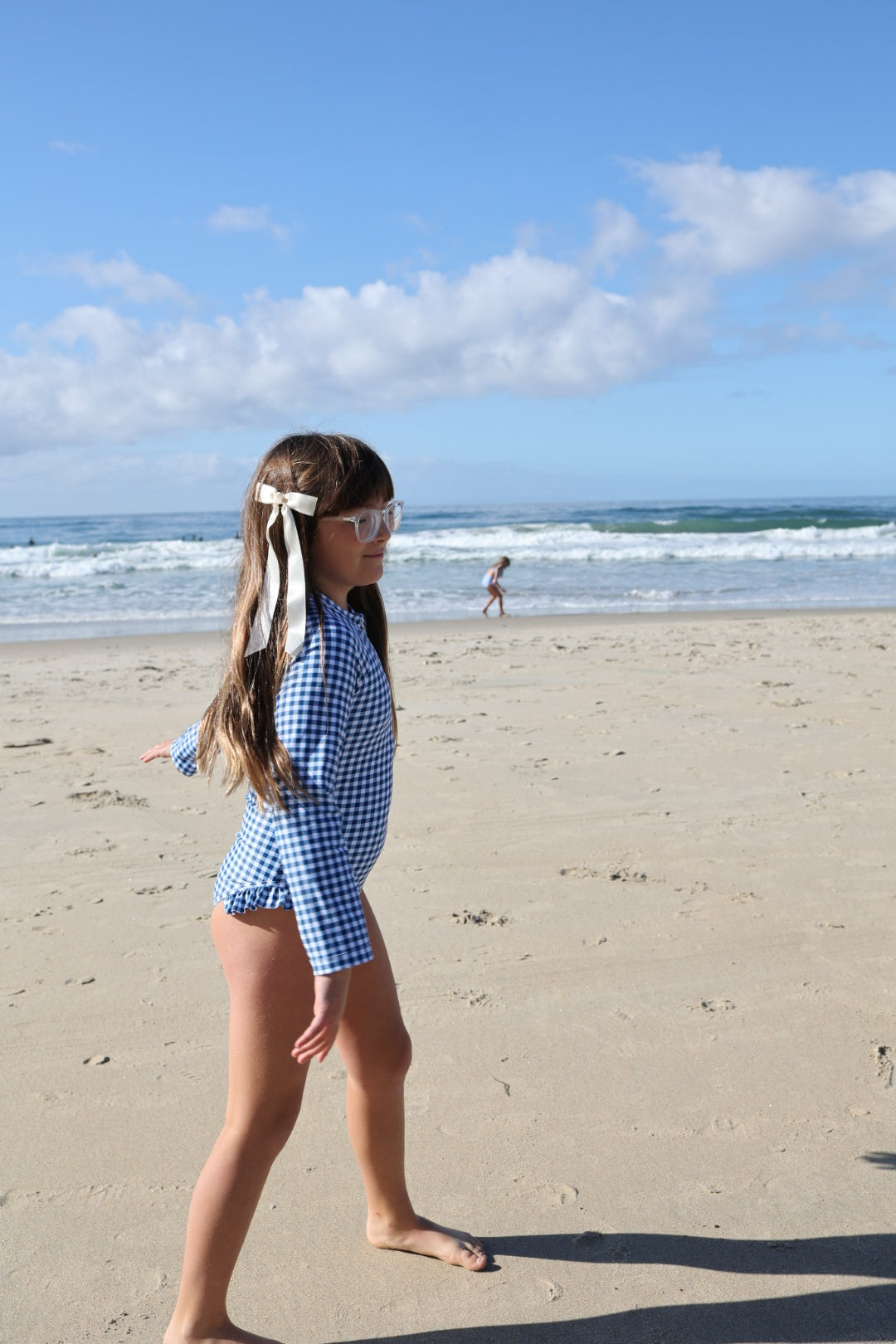A girl wearing the forever french baby Girls Long Sleeve Swimsuit in Blue Gingham, with white ribbons in her hair, walks along a sandy beach. The ocean and waves are behind her as another child plays near the water under a blue sky.