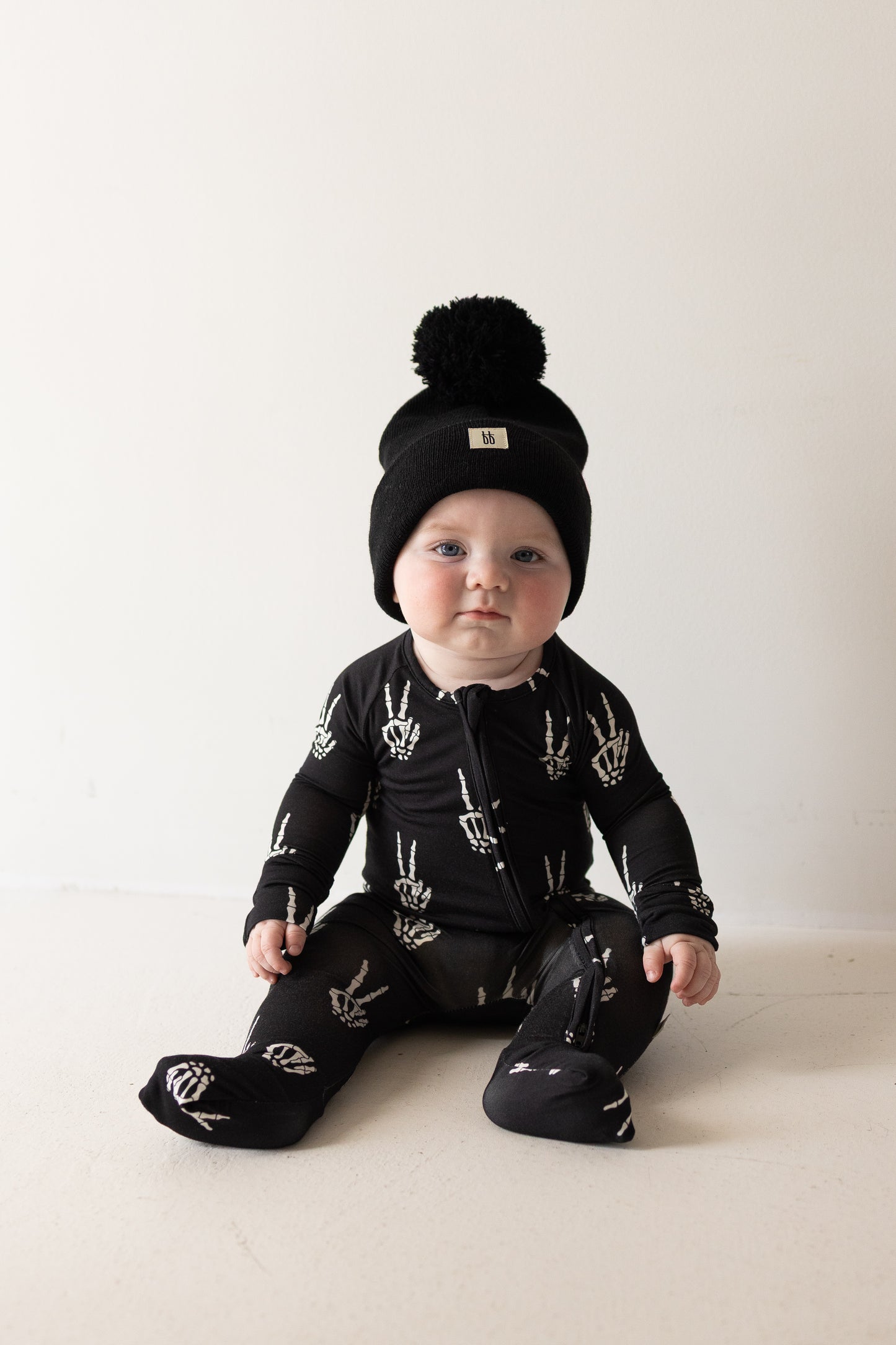 A baby dressed in the forever french baby Bamboo Zip Pajamas | Midnight Skelly—crafted from soft, hypo-allergenic bamboo viscose and featuring skeleton handprints—paired with a black pom-pom beanie, sits on a light floor against a plain white background, showcasing stylish comfort.