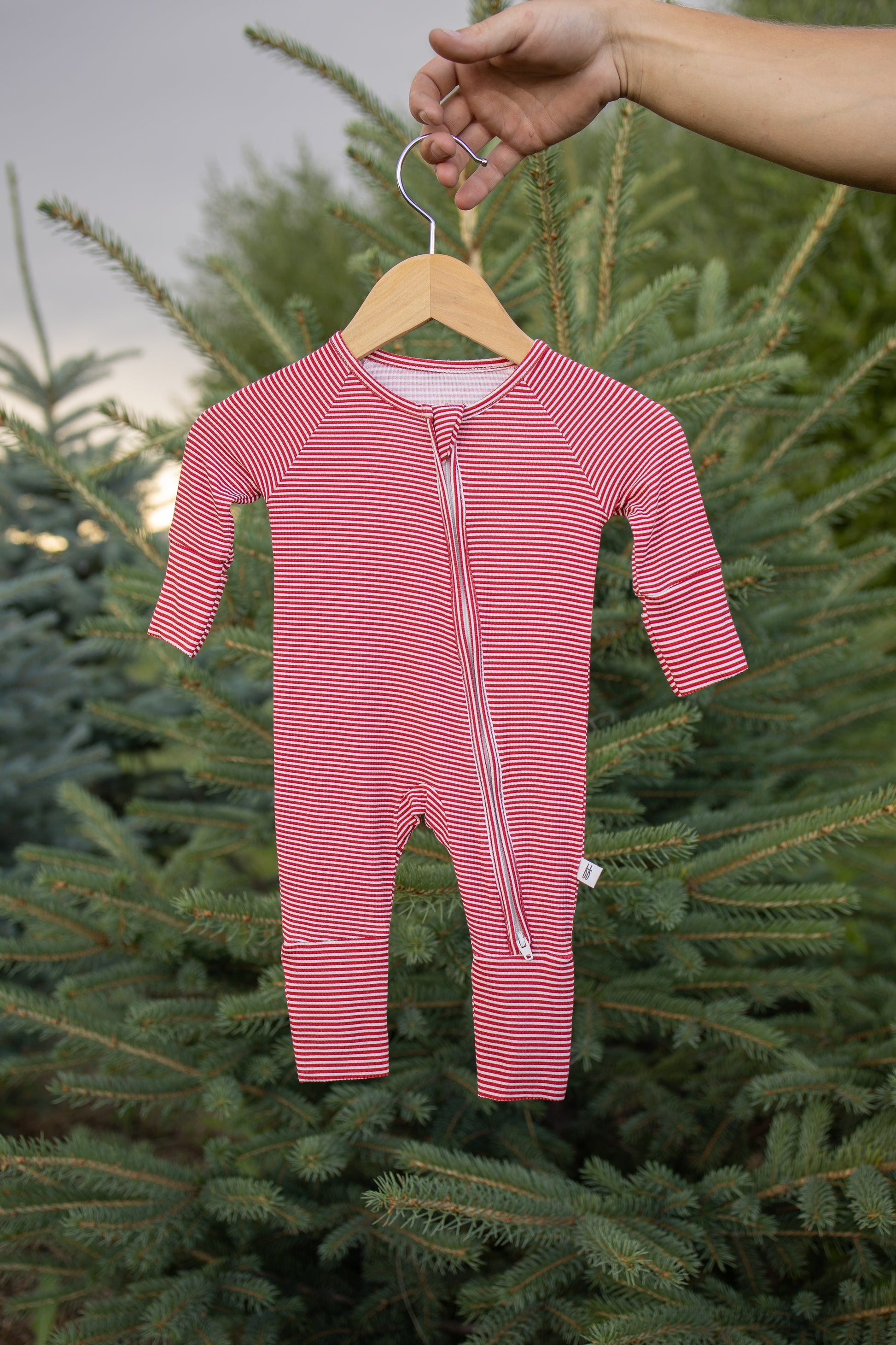 A hand holds a hanger displaying the forever french baby Bamboo Zip Pajamas in Peppermint Stripe Ribbed in front of green pine trees outdoors.