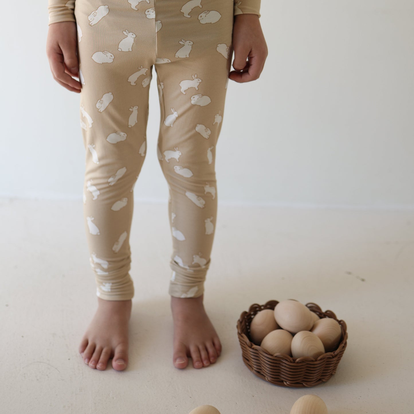 A child in forever french baby Bamboo Two Piece Pajamas | Bunnies stands barefoot beside a brown basket of wooden eggs on a light-colored floor.