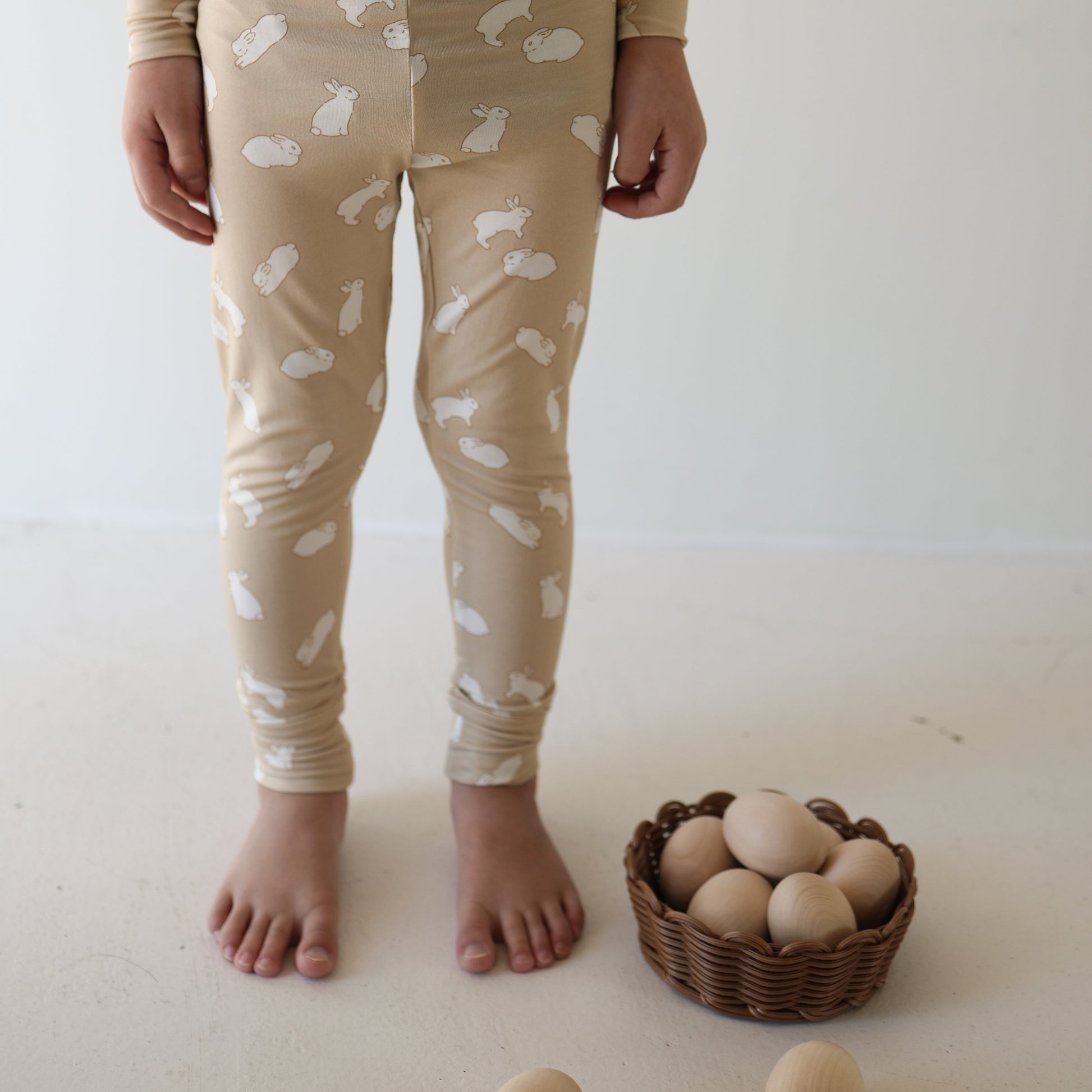 A child in forever french baby Bamboo Two Piece Pajamas | Bunnies stands barefoot beside a brown basket of wooden eggs on a light-colored floor.