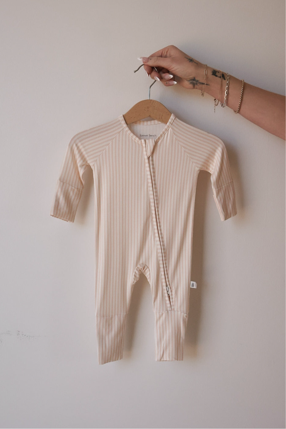 A hand holds a hanger with the forever french baby Bamboo Zip Pajamas in Sadie Stripe—a pale pink, long-sleeved, striped baby onesie featuring a diagonal zipper—shown against a plain light background.