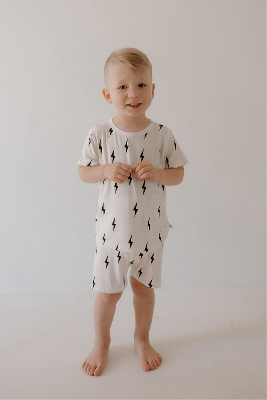 A young child with light hair smiles barefoot against a plain background, wearing the forever french baby Bamboo Short Set in White & Black Lightning Bolt—a breathable sleepwear t-shirt and shorts.