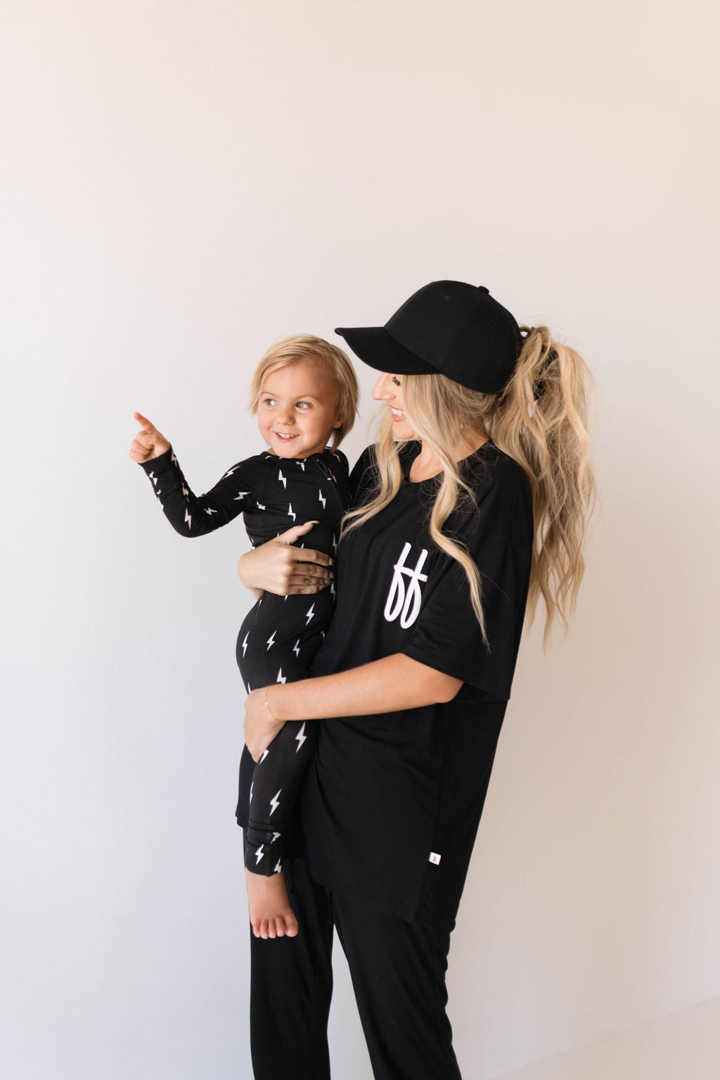 A woman with long blonde hair in a ponytail, dressed in the forever french baby Short Sleeve Women's Bamboo Lounge Set in Black ff Signature and a matching black cap, holds a smiling toddler. The toddler is wearing breathable bamboo fabric adorned with white lightning bolt patterns and is pointing with their right hand. Both are posed against a plain white background.