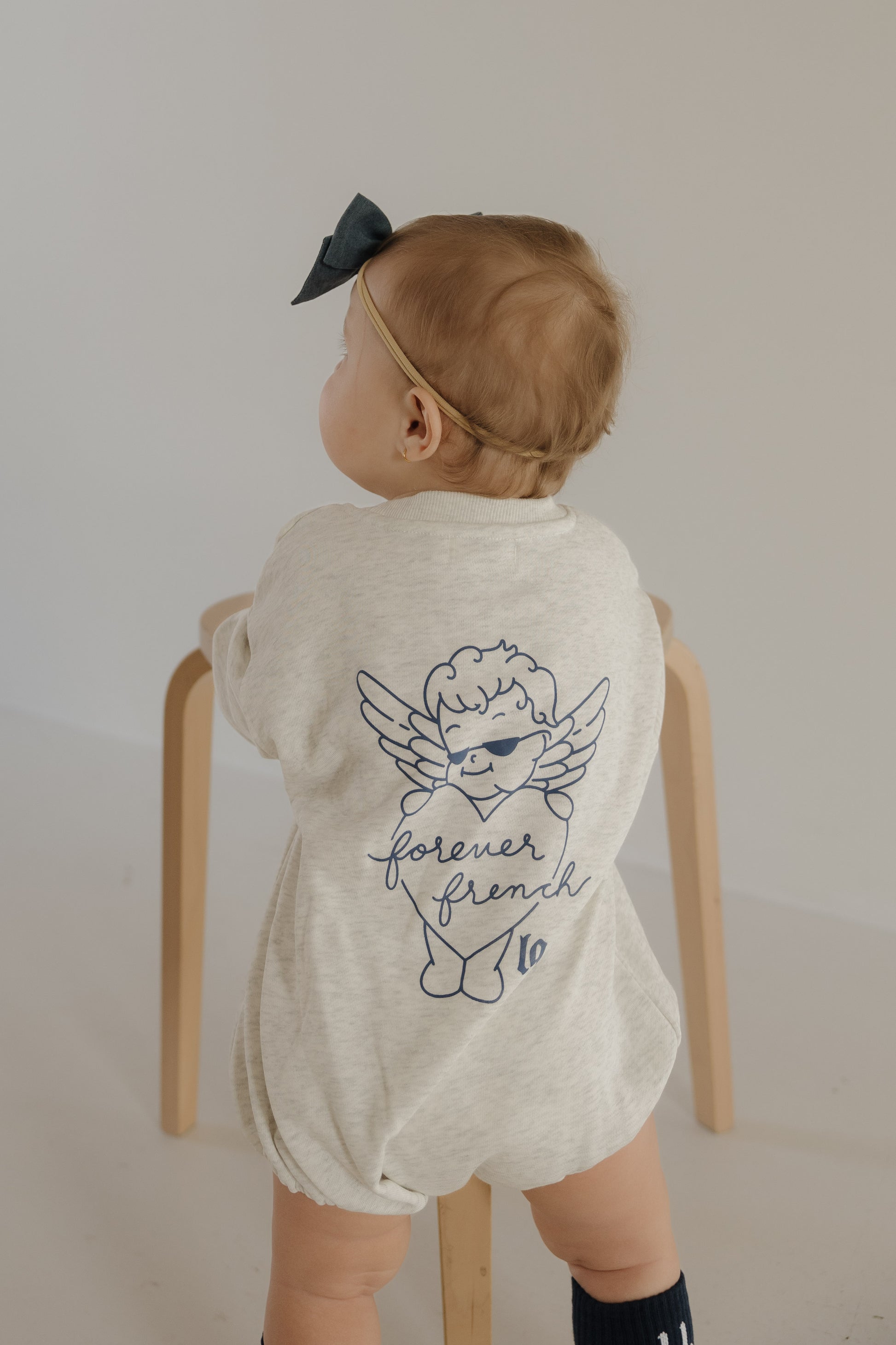 A baby wearing the "Sweatshirt Romper | ff Los Angeles" by forever french baby, in cream with an angel graphic and "forever french" on the back, stands holding a wooden stool, facing away from the camera.