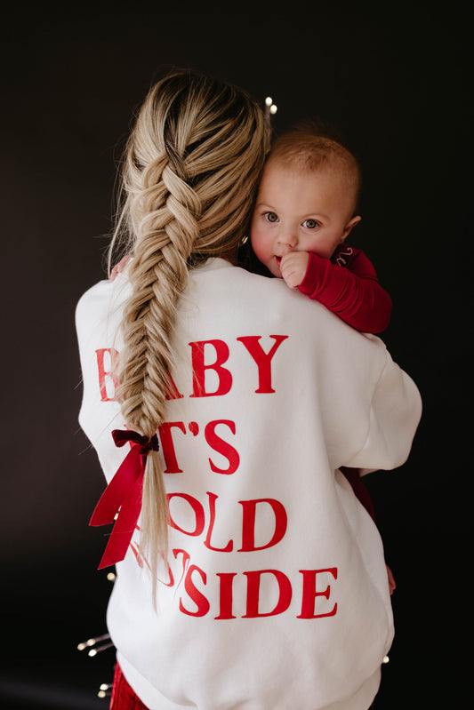 A woman with a long braid holds a baby dressed in red, capturing the holiday season spirit. She sports a chic white sweatshirt decorated with red letters that say, "Baby It's Cold Outside," from Forever French Baby's cozy collection. The scene is set against a dark backdrop brightened by festive lights, making it an ideal setting for the brand's offerings.