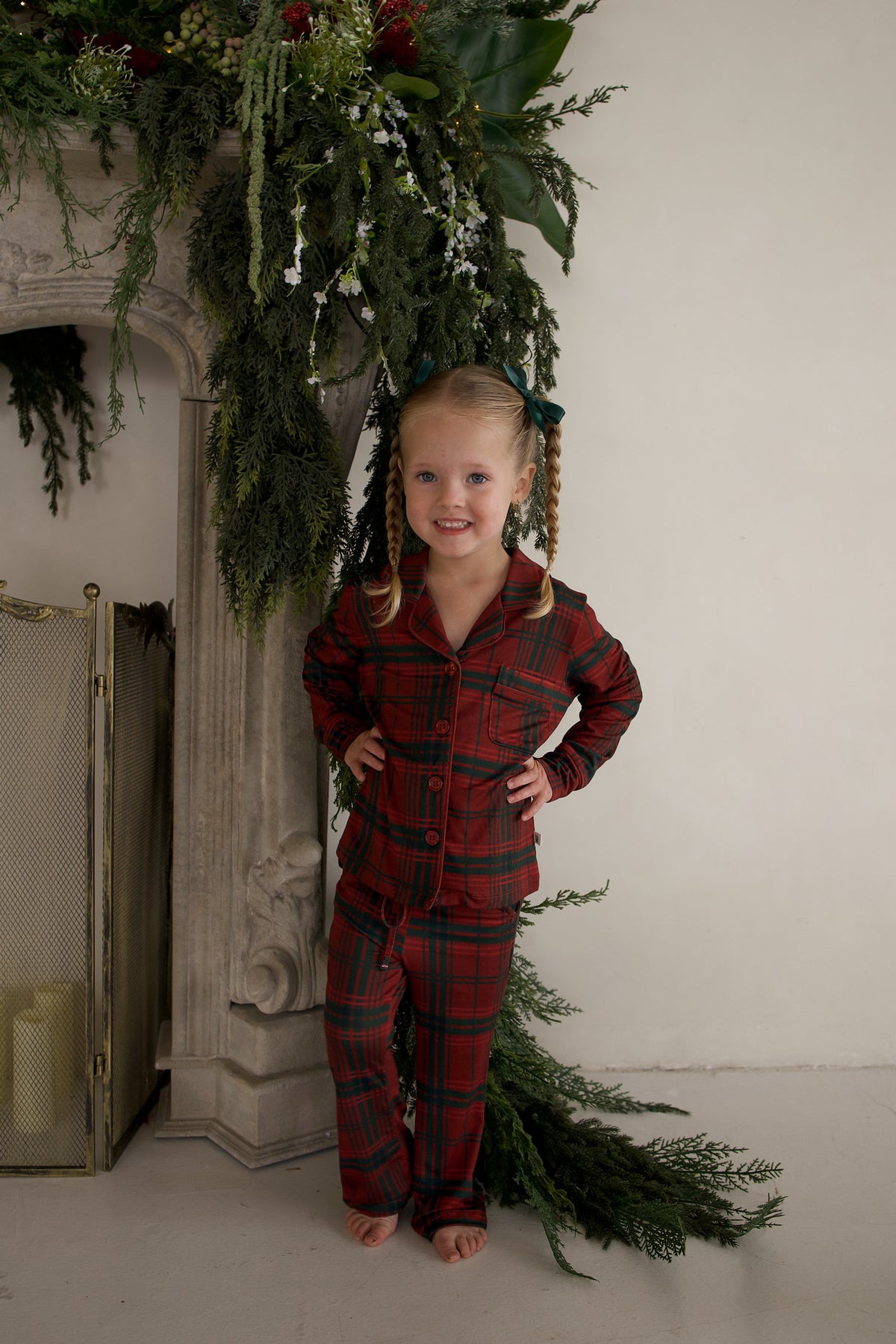 A young girl wearing a forever french baby Girl’s Bamboo Button Up Lounge Set in Christmas Plaid poses in front of a fireplace decorated with greenery.