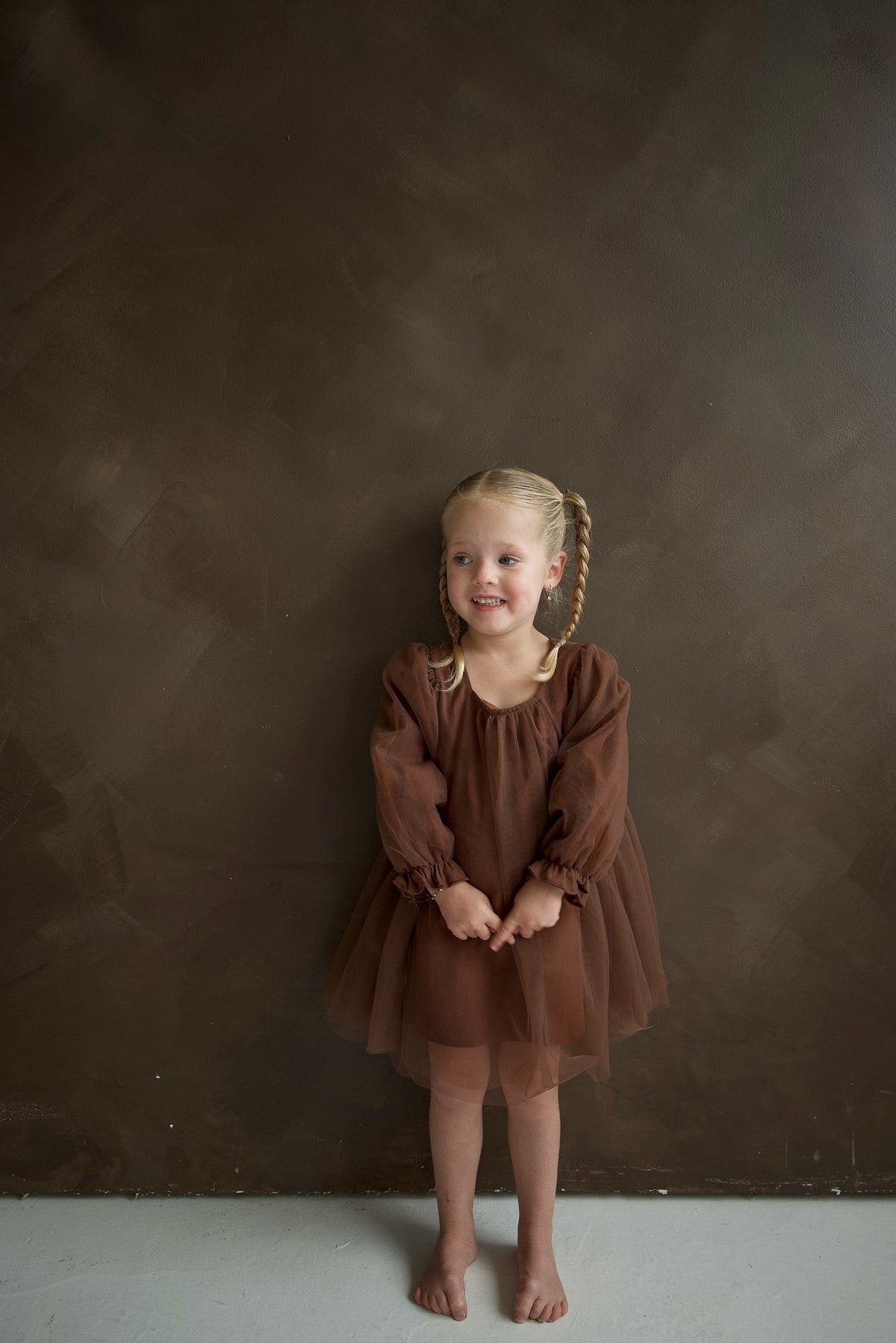 A young girl wears a brown Long Sleeve Tutu Dress | Cocoa while standing against a dark brown wall.