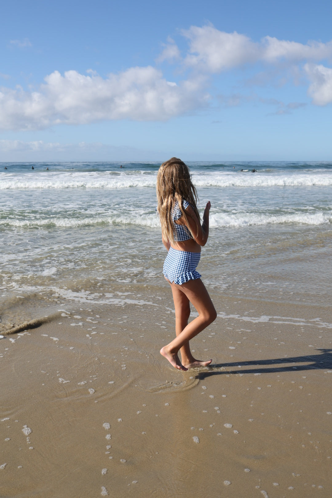 A young girl in the forever french baby Girls Two Piece Swimsuit | Blue Gingham walks barefoot along the wet beach sand, gazing at ocean waves beneath a bright, partly cloudy sky.