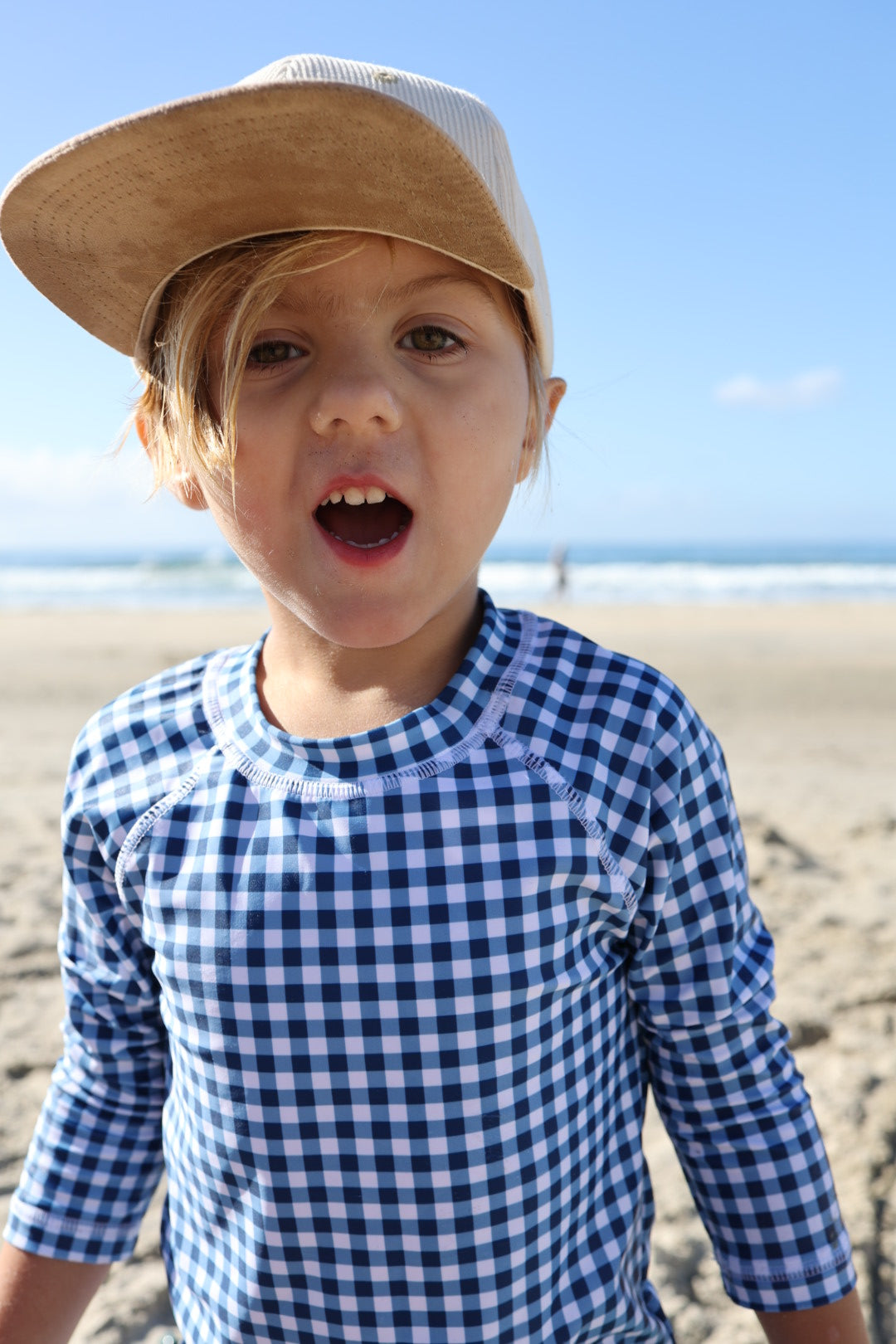 A young child wearing the forever french baby Child Rash Guard | Blue Gingham and a tan baseball cap stands on a sandy beach with the ocean and blue sky behind them, mouth open as if speaking or shouting.