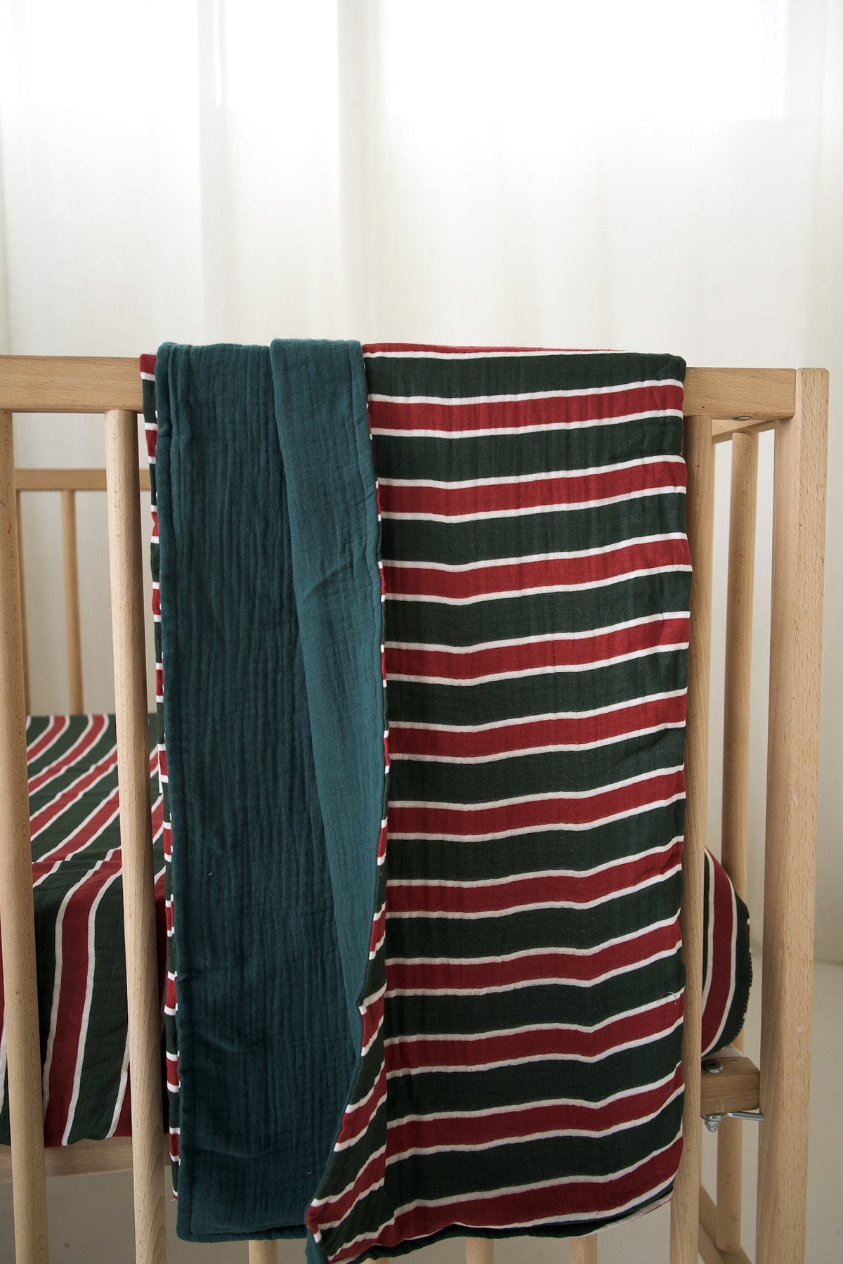 A forever french baby Muslin Quilt | Holiday Stripe with a red, white, and dark green striped pattern draped over a wooden crib.