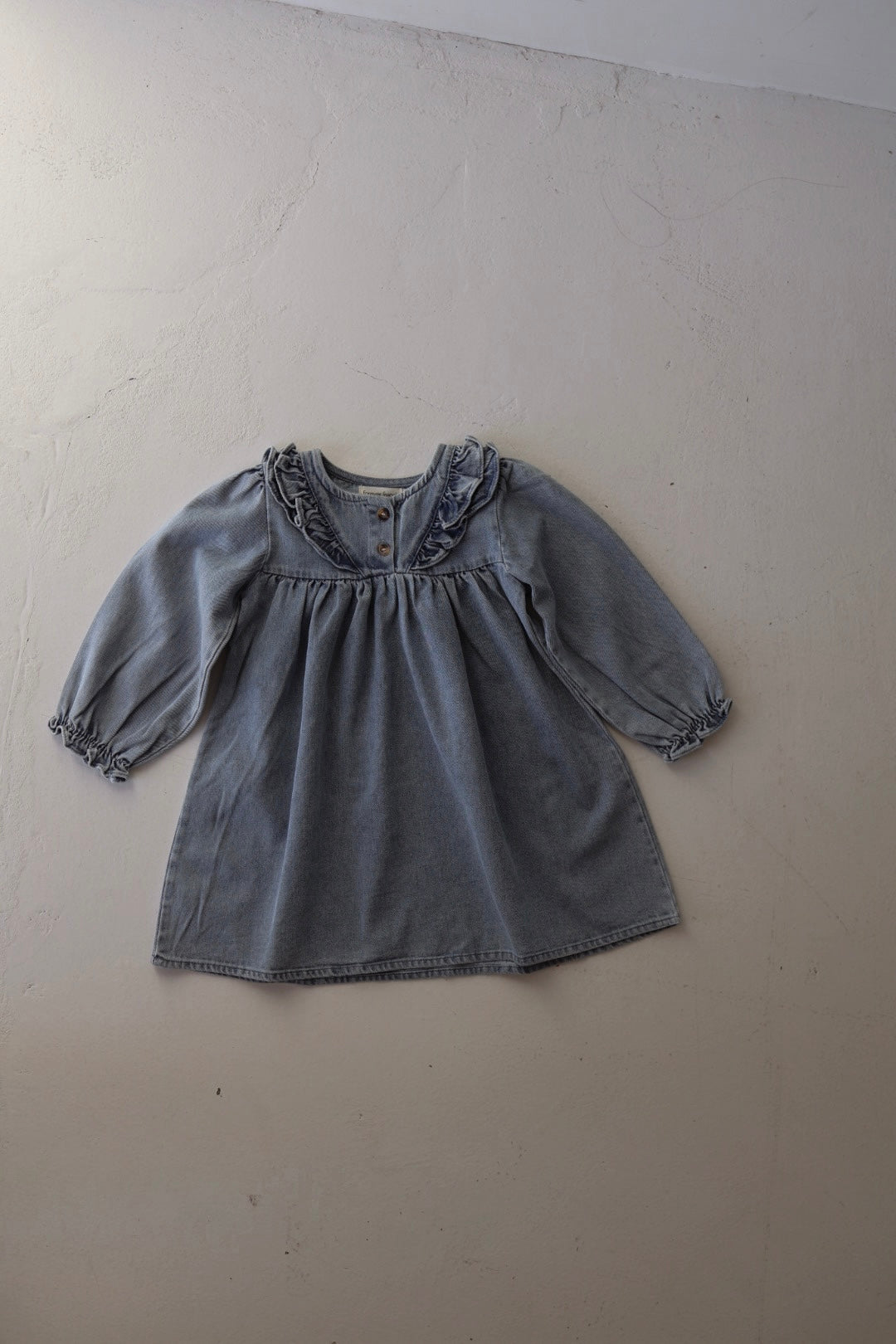 The forever french baby Denim Dress | Ruffle, a tunic-style long sleeve girls dress, is displayed flat on a light-textured surface. It features ruffled detailing at the collar and elasticated sleeve cuffs.
