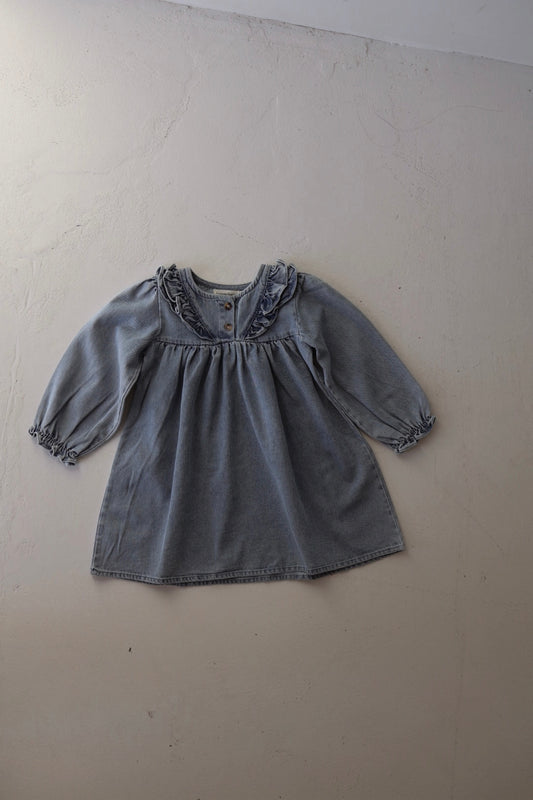The forever french baby Denim Dress | Ruffle, a tunic-style long sleeve girls dress, is displayed flat on a light-textured surface. It features ruffled detailing at the collar and elasticated sleeve cuffs.