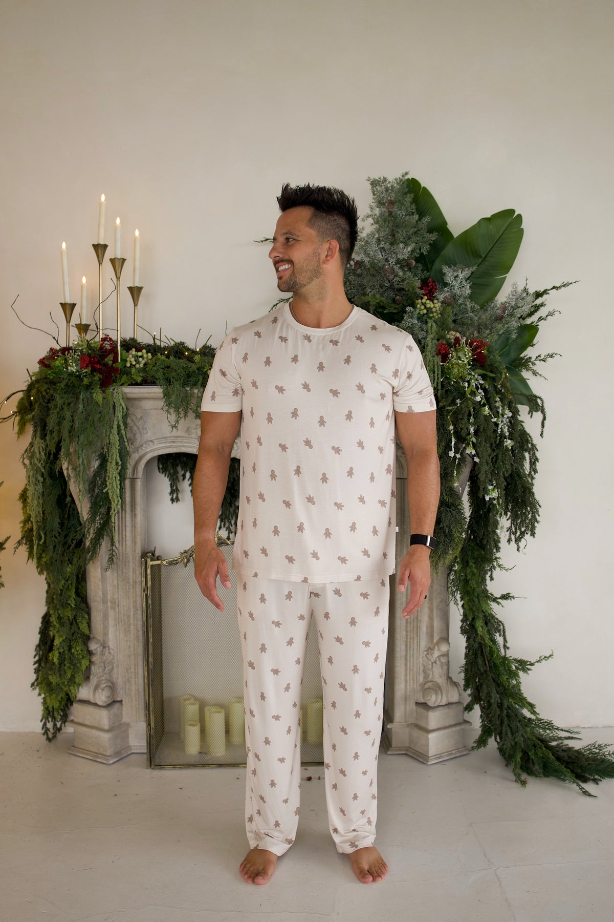 A man wearing a Men’s Short Sleeve Pajamas | Gingersnap set with a repeating gingerbread man pattern, standing in front of a fireplace decorated with greenery and candles.
