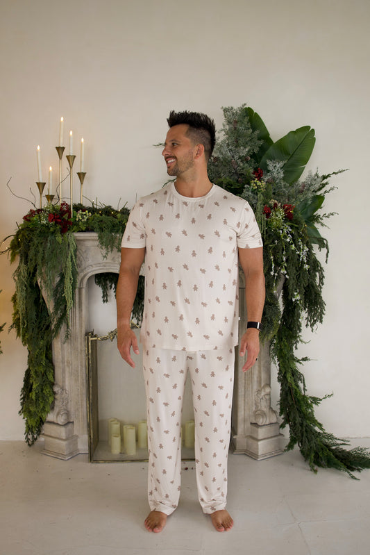A man wearing a Men’s Short Sleeve Pajamas | Gingersnap set with a repeating gingerbread man pattern, standing in front of a fireplace decorated with greenery and candles.