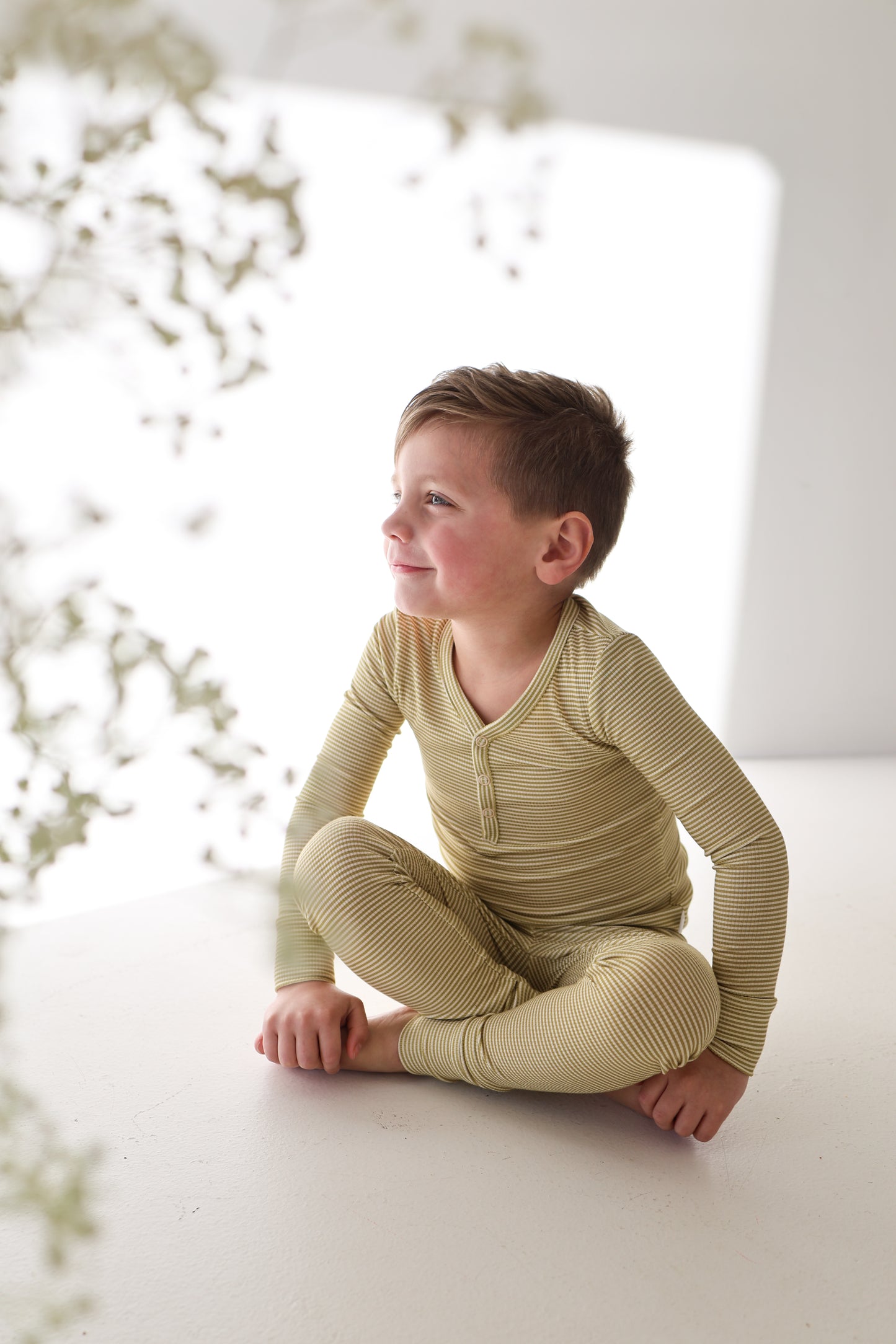 A young boy with short brown hair wears forever french baby Bamboo Two Piece Pajamas in Willow Stripe Ribbed, sitting cross-legged and smiling in a bright, minimalistic room filled with soft natural light.