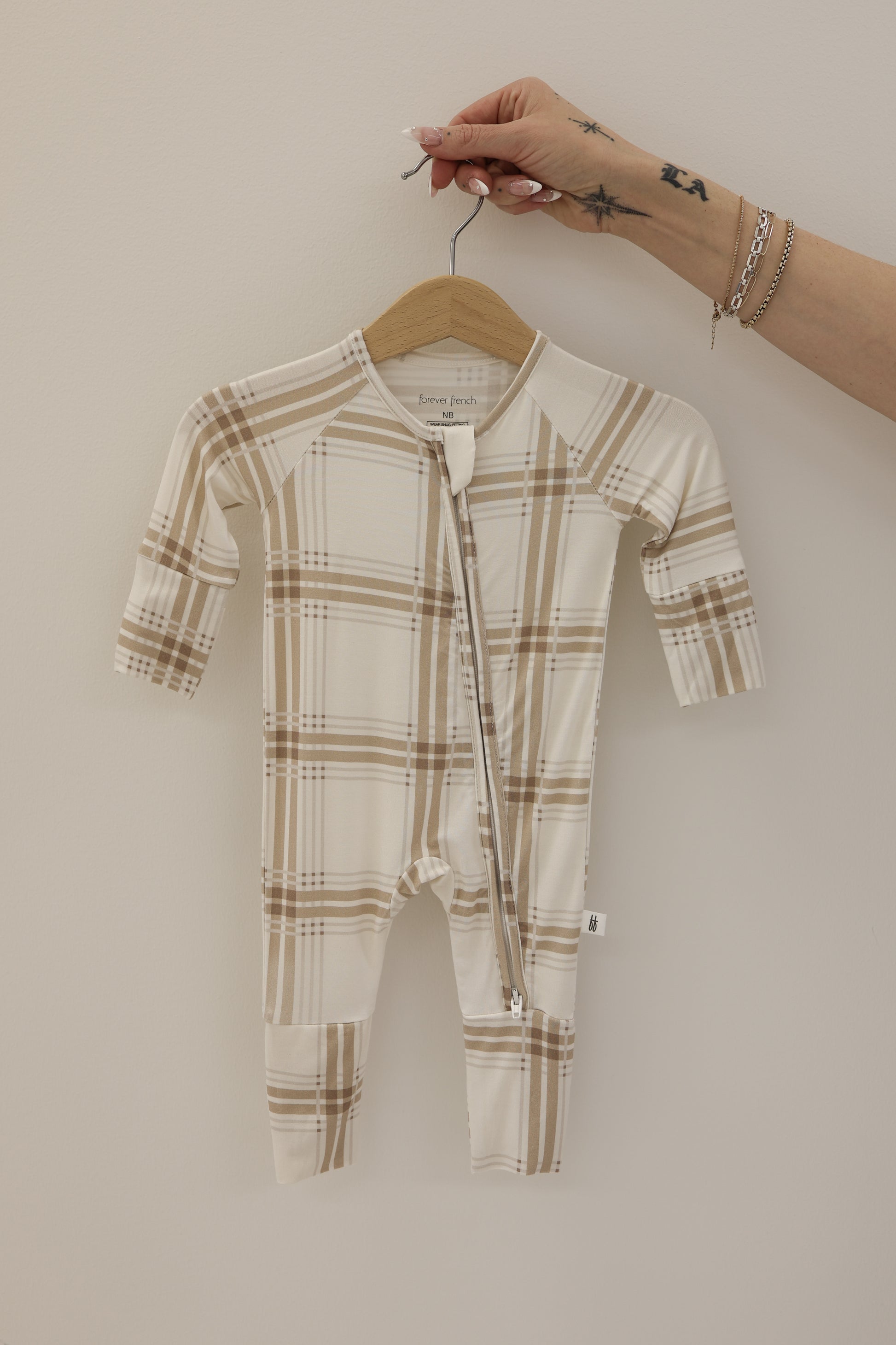 A hand with star tattoos holds a wooden hanger displaying the forever french baby Bamboo Zip Pajamas in Ellis Plaid—beige and white, hypoallergenic, with a diagonal zipper—against a plain light background.