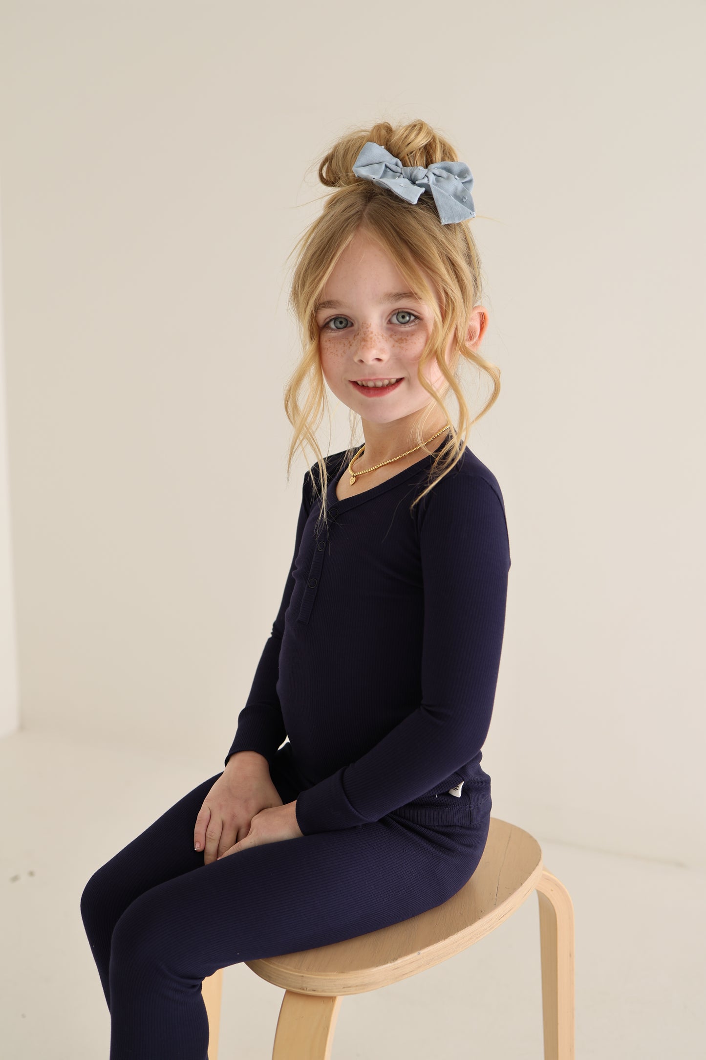 A young girl with light skin and freckles, smiling at the camera, wears forever french baby Bamboo Two Piece Pajamas in Blueberry Ribbed and a large blue bow, sitting on a wooden stool against a plain light background in comfy, hypoallergenic kids clothing.