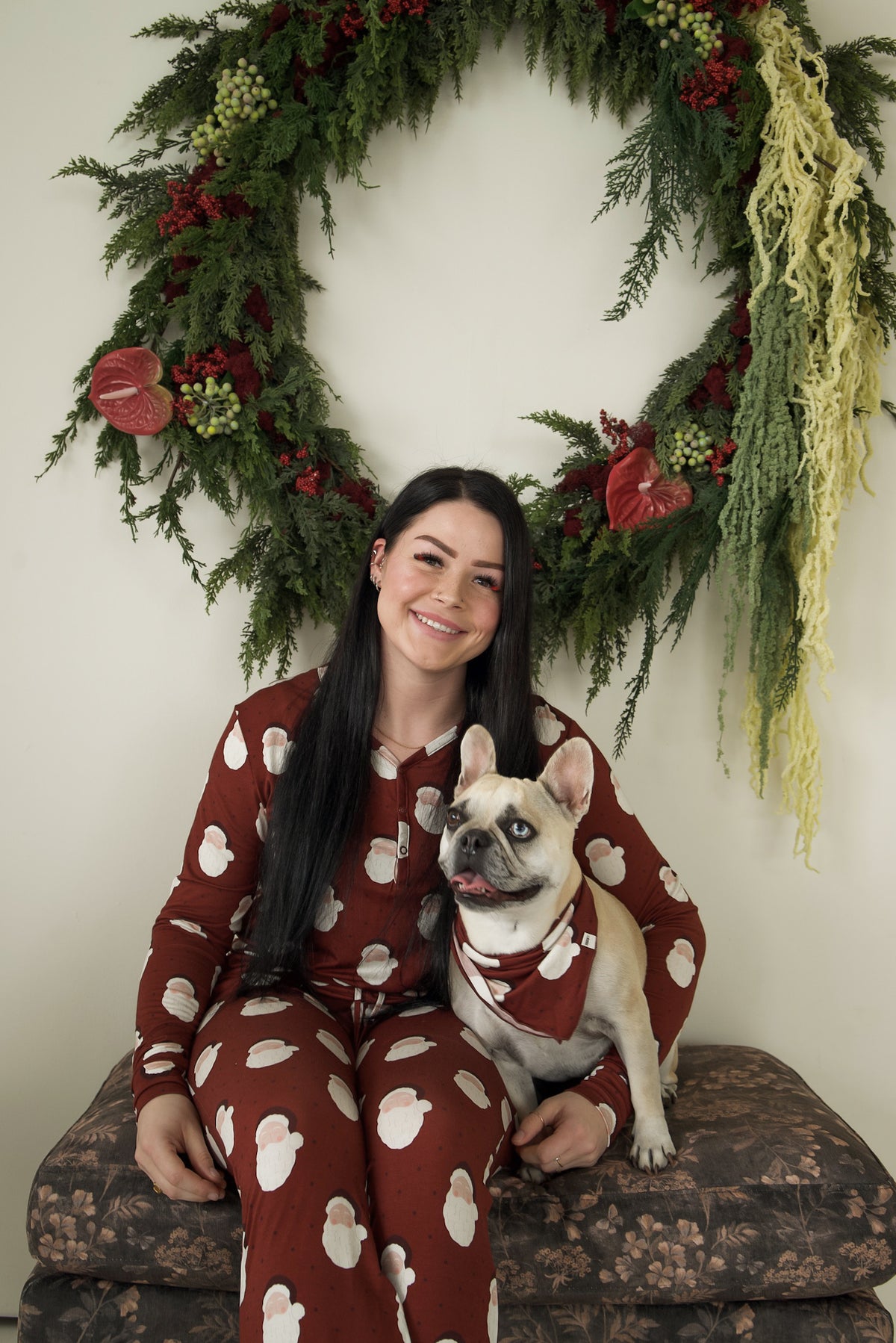 A woman and a French Bulldog wearing a Bamboo Bandana | Vintage Santa sit in matching burgundy vintage Santa-print bamboo viscose outfits in front of a large Christmas wreath.