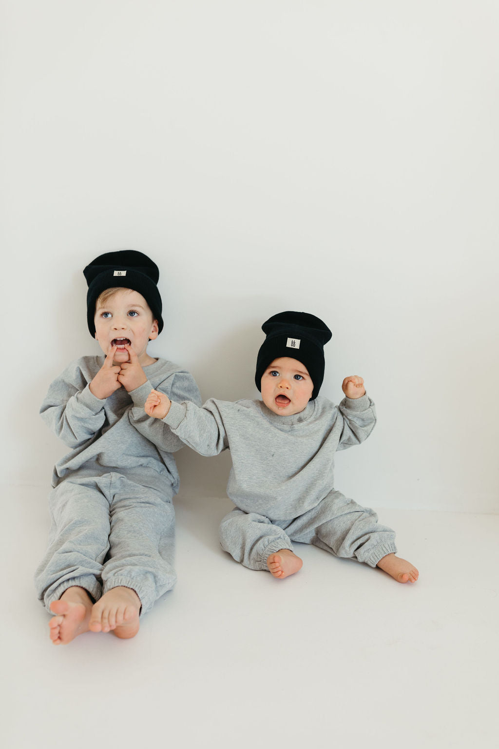 Two young children sit on the floor against a plain white background, both dressed in matching "Stormi" sweat sets by forever french baby and wearing black beanies. The older child sits with their mouth open, while the younger one raises both hands with a curious expression.