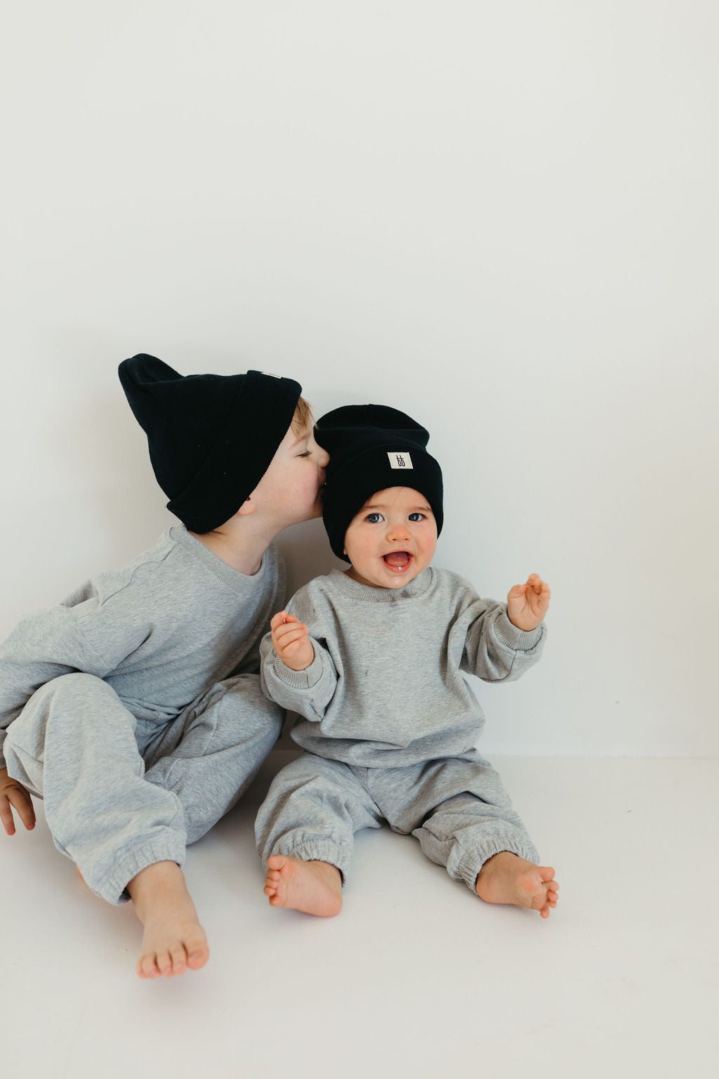 Two toddlers dressed in matching 'Stormi' sweat sets by forever french baby and sporting black beanies are seated on the floor. One child leans in for a kiss while the other, smiling and holding a pacifier, reciprocates. They are barefoot against a simple, light backdrop.