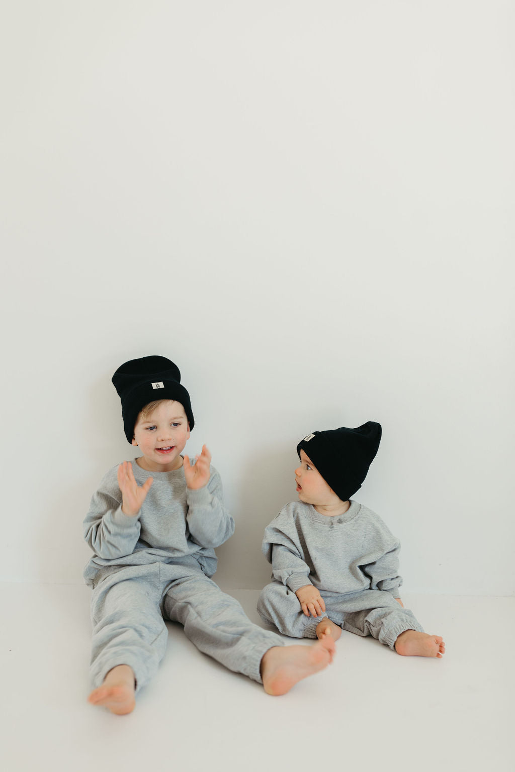 Two young children sit against a plain white background, both dressed in matching "Sweat Sets | Stormi" by forever french baby and wearing black beanies. The older child claps while the younger looks on. They are barefoot and appear delighted, their joy contagious in this simple yet charming scene.