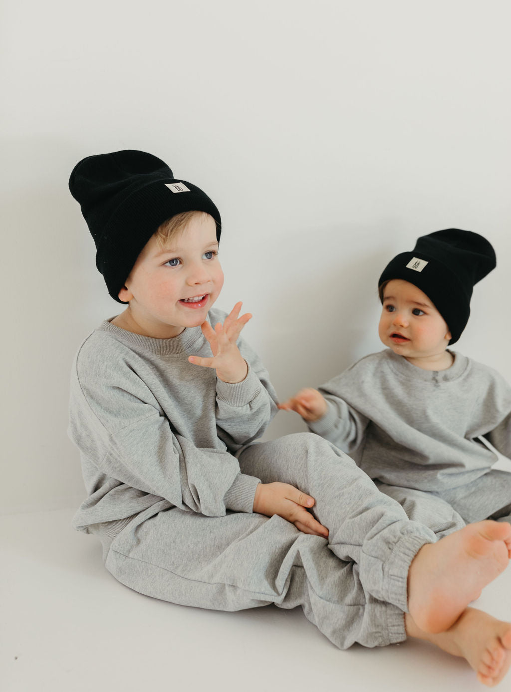 Two young children sit together against a plain background, dressed in matching Stormi sweat sets by forever french baby, complemented with black beanies. One child, wearing cuffed joggers from the set, smiles while looking to the side, as the other glances at them with curiosity.