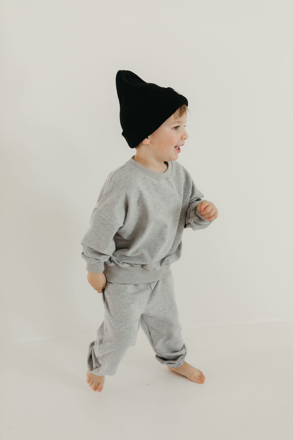 A young child in the forever french baby "Sweat Set | Stormi," which includes a grey crewneck sweater and cuffed joggers, poses upright with a playful expression against a plain white background, wearing a stylish black beanie.