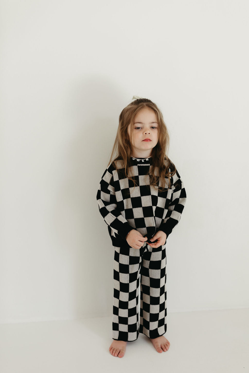 A young girl stands barefoot against a simple wall, dressed in the Knit Pant Set by forever french baby, featuring a black checkerboard design with long sleeves and cozy knit pants. Her hands are clasped in front of her as her long hair softly drapes around her shoulders.