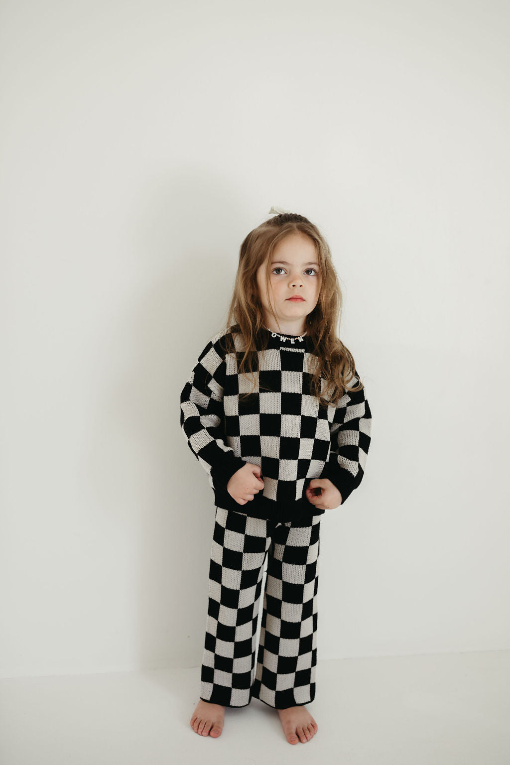 A young child stands barefoot against a plain white background, dressed in the Knit Pant Set | Black Checkerboard from forever french baby. The oversized sweater drapes gently as the child's long hair flows down, giving them a serious expression.