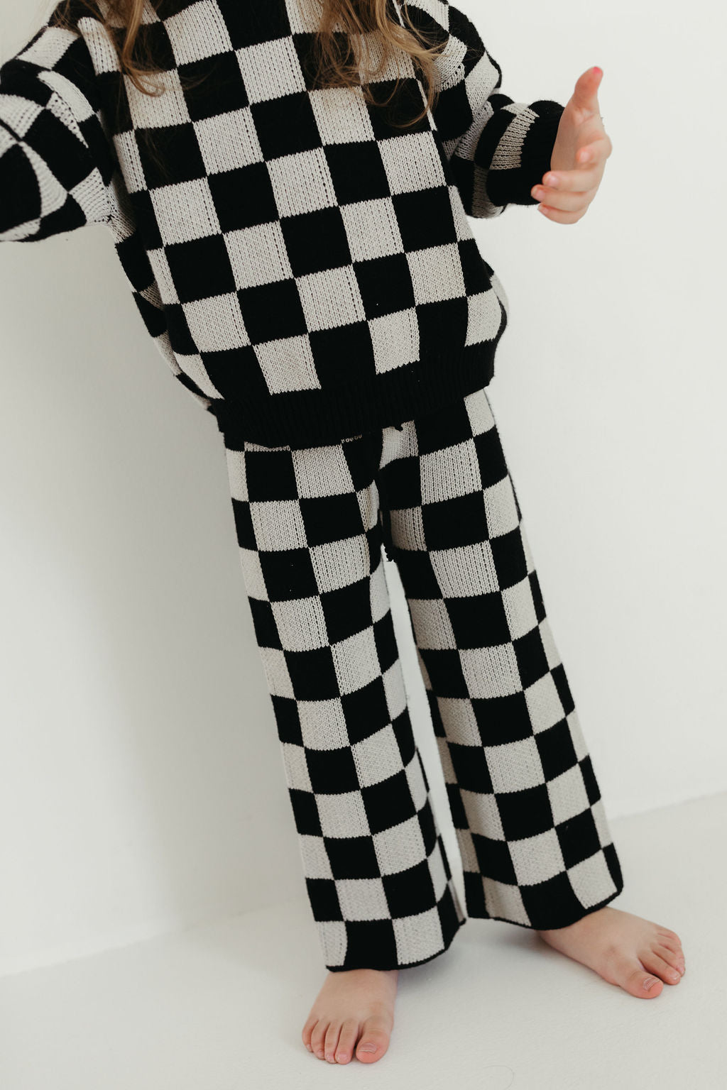 A child wearing the "Knit Pant Set | Black Checkerboard" from forever french baby, which includes an oversized sweater and comfy knit pants, stands barefoot on a white floor. Only the lower half of the child's face is visible, while the bold geometric pattern offers a striking contrast.