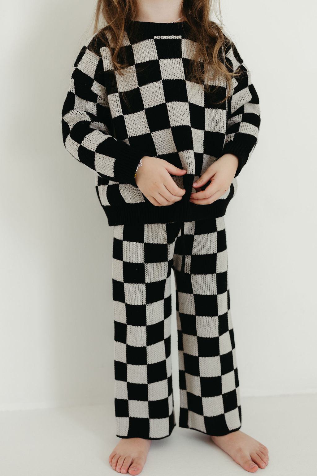 A child is dressed in the Knit Pant Set | Black Checkerboard from forever french baby, featuring comfortable knit pants and a long-sleeved top, standing against a white backdrop. The cozy design of the outfit is highlighted as the child's hands rest near their waist with bare feet peeking into view, while their face remains unseen.