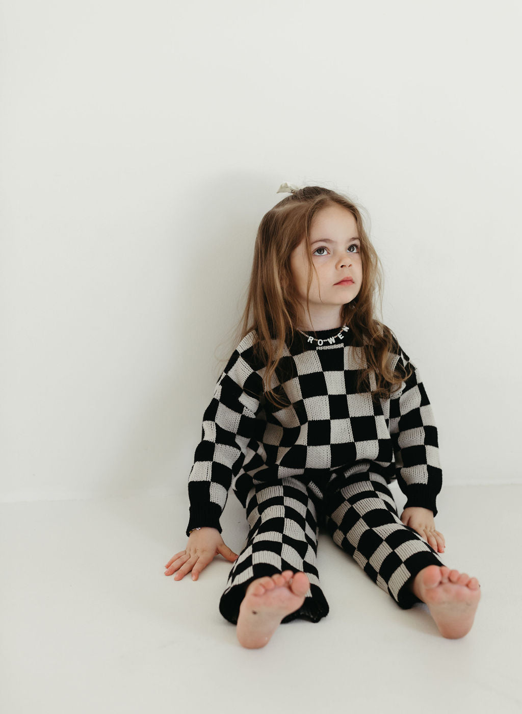 A young child with long hair sits barefoot against a plain white wall, clad in the forever french baby Knit Pant Set in Black Checkerboard. The cozy knit pants enhance their upward gaze, contributing to the scene's serene simplicity.