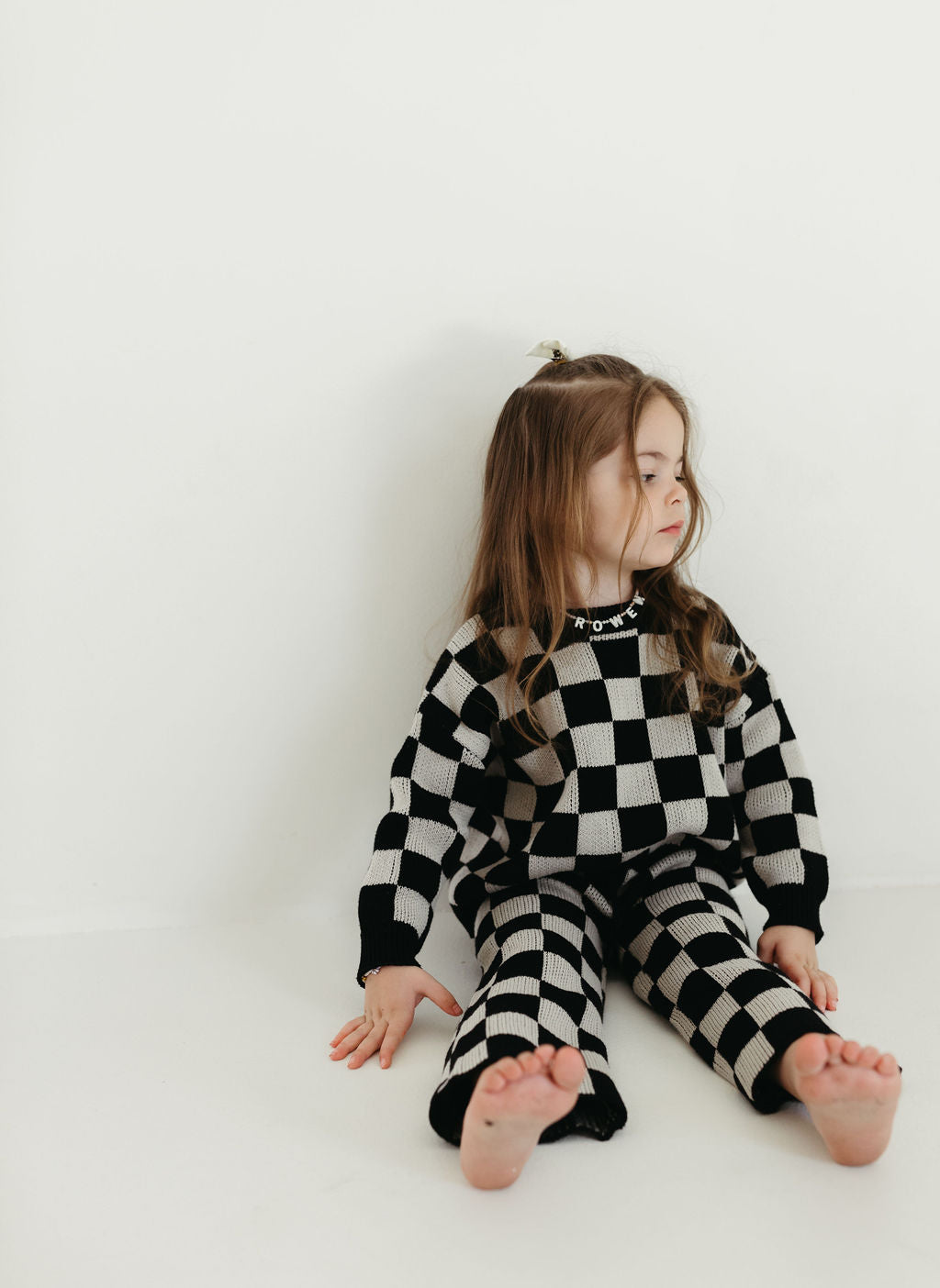 A young girl with long hair sits on the floor against a white wall, dressed in the Knit Pant Set | Black Checkerboard by forever french baby. She appears to be looking off to the side with a thoughtful expression.