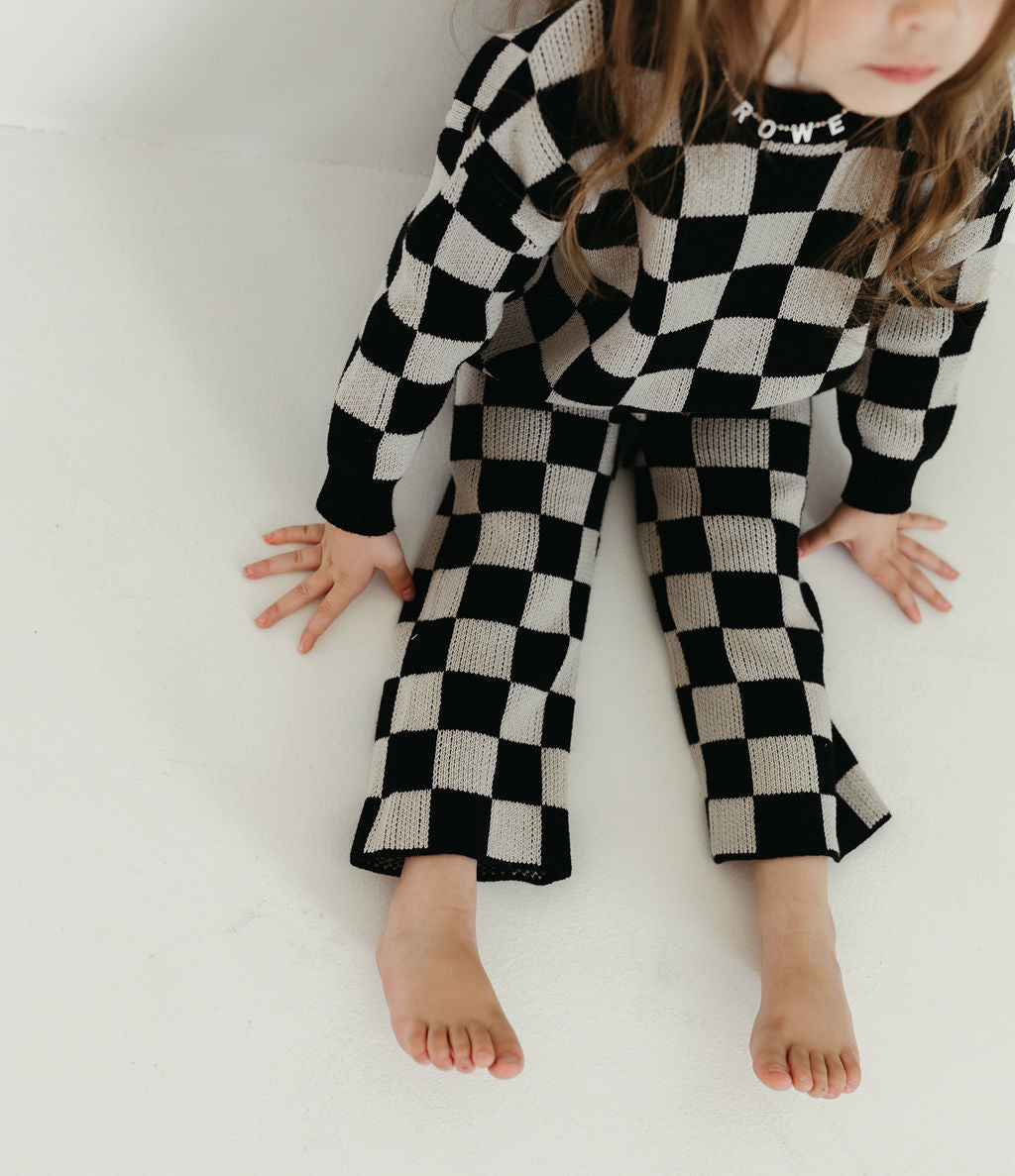 A child wearing the "Knit Pant Set | Black Checkerboard" by forever french baby sits on the floor barefoot. The oversized sweater displays a partially visible word, set against a plain white background.