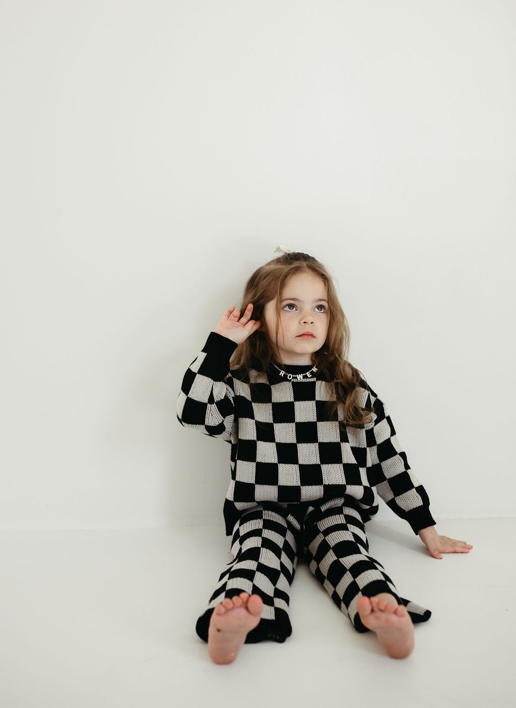 A young girl sits against a light wall, enveloped in the Knit Pant Set | Black Checkerboard by forever french baby. Her hair flows freely as she gazes upward with a thoughtful expression, resting one hand near her ear.