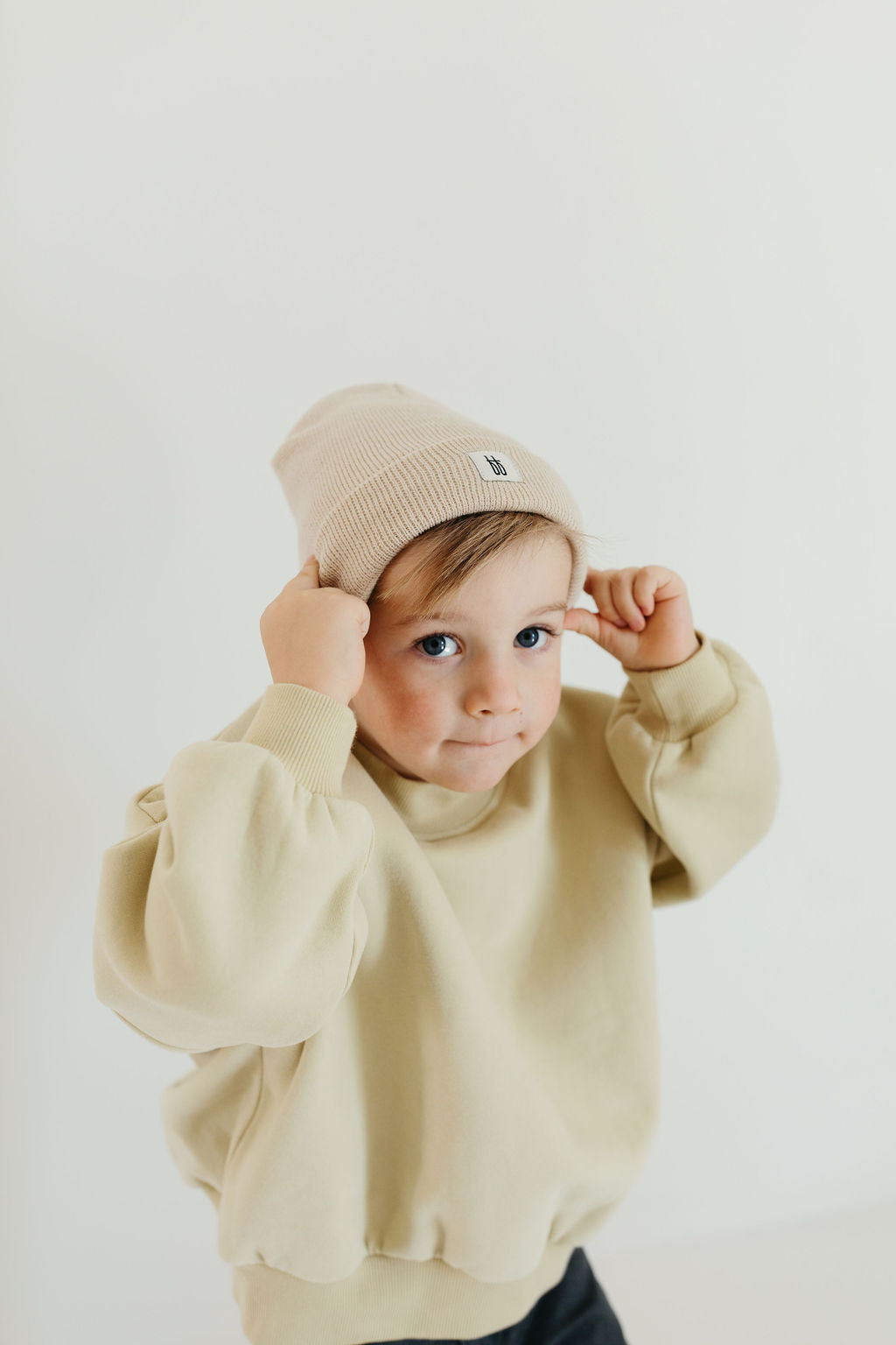 A child dressed in the Child Sweat Set from forever french baby, which includes a beige sweater and matching beanie, gazes at the camera with a neutral expression. The plain white background enhances the soft and minimalist composition.