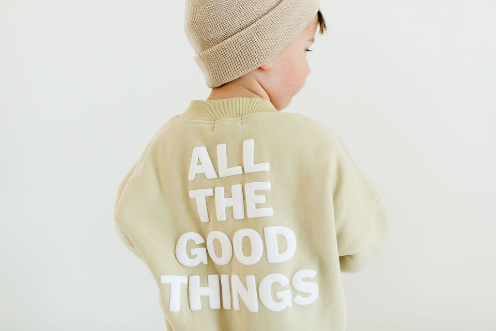 A child in a Forever French Baby ensemble stands against a white background, looking stylish and cozy in their beige beanie and cream-colored Child Sweat Set | All the Good Things.