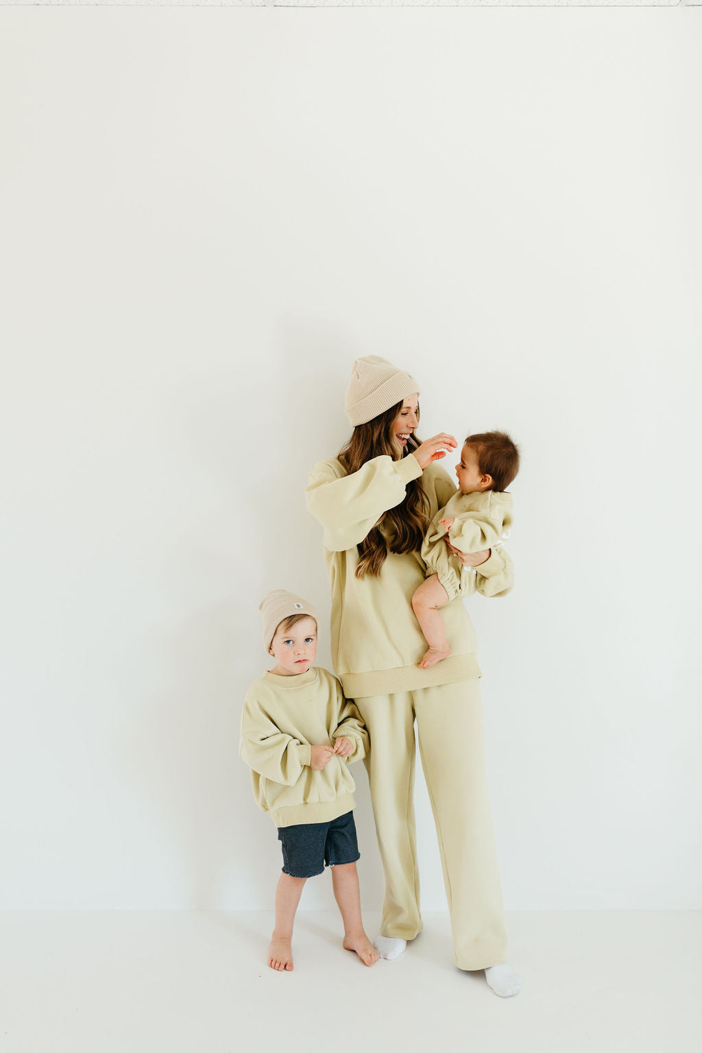 A woman dressed in a beige outfit and beanie hat holds a baby wearing a matching ensemble from Forever French Baby. Next to her, a young child stands out in an avocado green romper from the Child Sweat Set - All the Good Things collection, perfectly complementing the neutral tones against a plain white background.