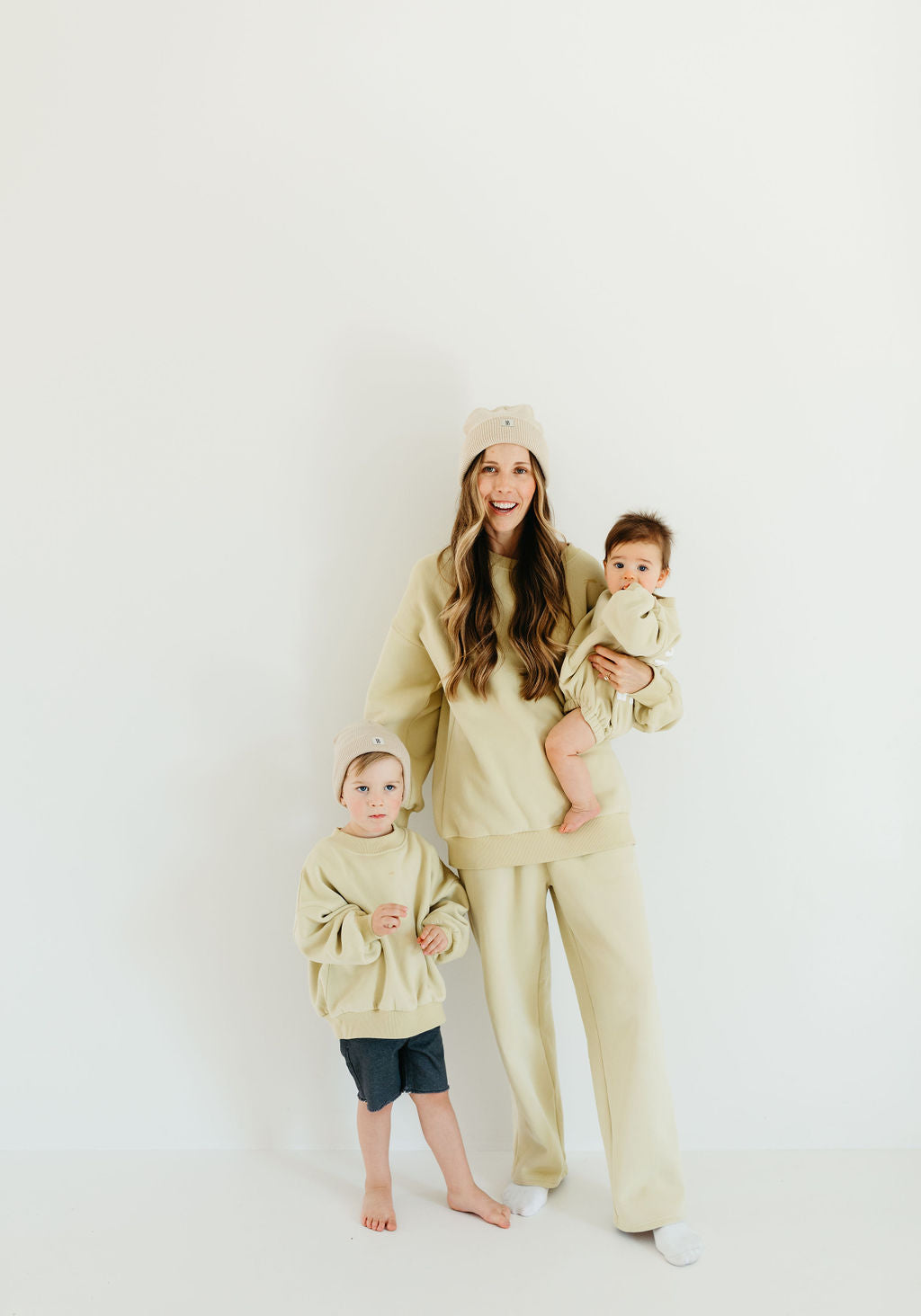 A woman poses against a white backdrop in matching beige sweat sets alongside two young children. One child is standing, while the other is held by the woman. All three appear content and coordinated in their cozy Forever French Baby attire, specifically from the "Child Sweat Set | All the Good Things" collection.