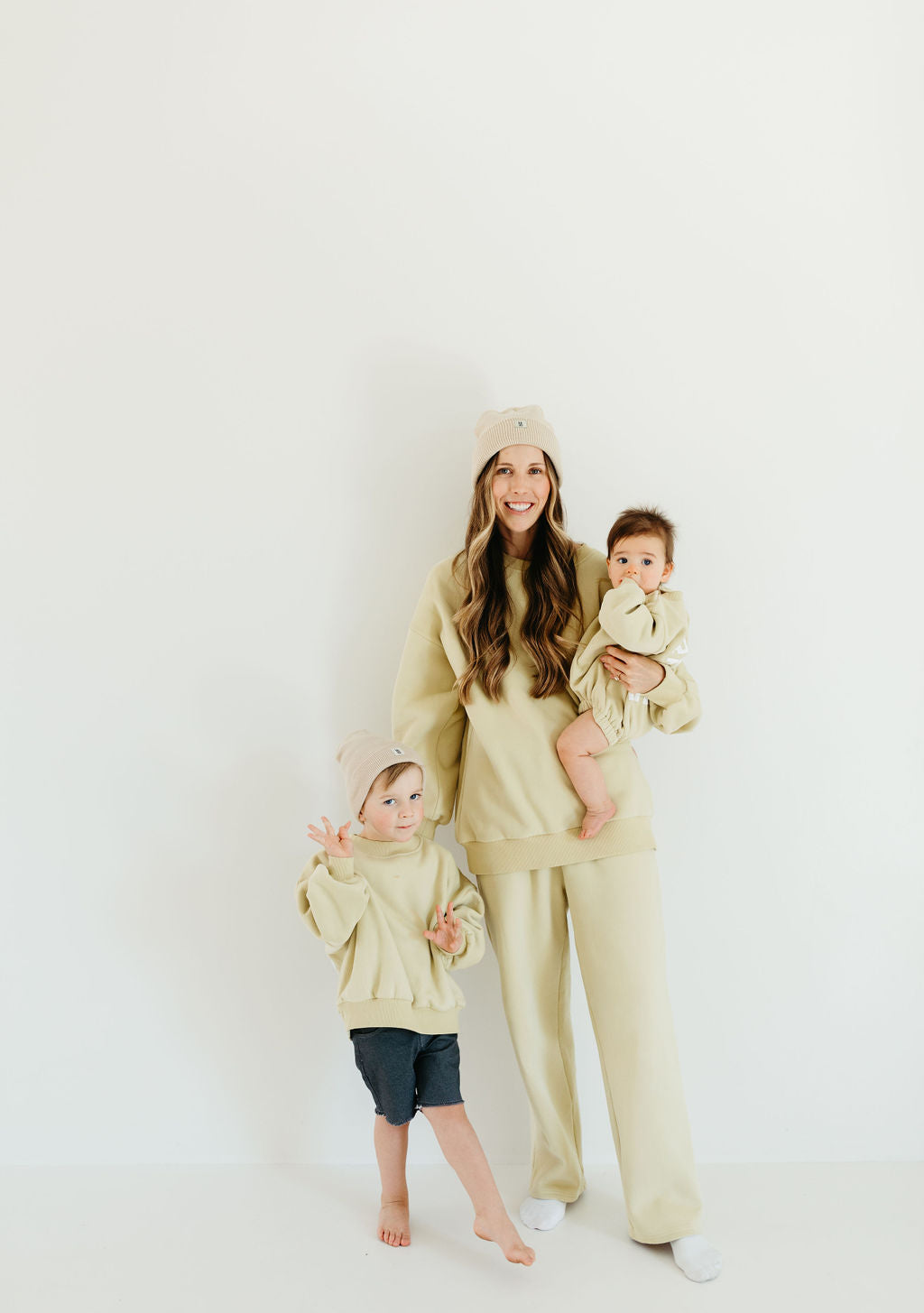 A woman stands against a white background, wearing a matching cream-colored sweat set. She holds a baby in an outfit similar to the Child Sweat Set from All the Good Things by Forever French Baby and stands next to an older child in a cream sweatshirt and gray shorts. Both children wear beanies.