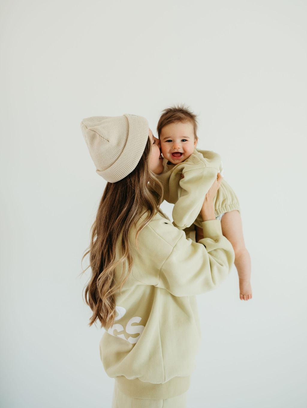 A person wearing a beige beanie and matching outfit holds and kisses a smiling baby dressed in the "All The Good Things" sweatshirt romper by forever french baby. The background is plain white.